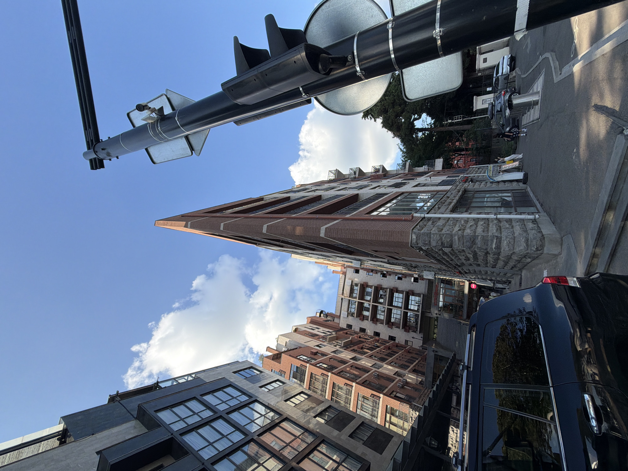 This media file captures a street scene in Tbilisi, Georgia. The perspective is low-angle, looking up towards a bright blue sky dotted with fluffy white clouds. In the foreground, a black vehicle, possibly an SUV or van, is parked on the right side of the road.
Dominating the scene are several multi-story buildings with a mix of architectural styles. The building closest to the foreground on the left appears to be modern, with a dark facade and large, grid-like windows. To its right, a prominent building with a distinctive triangular facade is covered in red brick and has many windows. Further back, other buildings showcase a variety of brickwork, balconies, and window arrangements.
Overhead, a traffic signal arm extends across the frame, equipped with traffic lights, signs, and a camera. The sunlight suggests it is daytime, likely mid-morning or afternoon, with clear weather. The presence of trees and the architectural style of the buildings provide location cues for Tbilisi. The overall impression is one of an urban environment on a pleasant day. There is no visible text in the image.