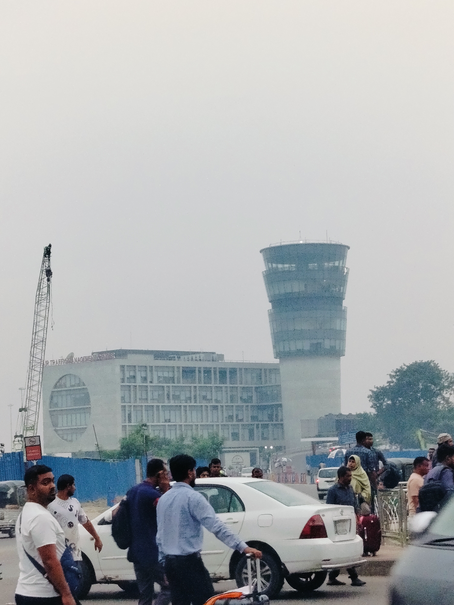 The image depicts an outdoor scene at Hazrat Shahjalal International Airport in Dhaka, Bangladesh. In the background, a modern airport control tower with a distinctive cylindrical shape and glass facade stands tall against a hazy sky. Adjacent to it is a large, multi-story building with numerous windows, likely an airport terminal or administrative building, and a large crane is visible to the left of this building, suggesting ongoing construction or maintenance.
In the foreground, a white sedan is parked on a paved area, and several people are visible around it. Some individuals are walking, while others appear to be waiting or interacting. One man in a white t-shirt is carrying a dark blue shoulder bag, and another man in a light blue shirt is walking towards the right of the frame. A woman wearing a light yellow hijab is standing with a suitcase. The overall atmosphere is busy, with several vehicles and pedestrians present, typical of an airport environment. The weather appears overcast or hazy.