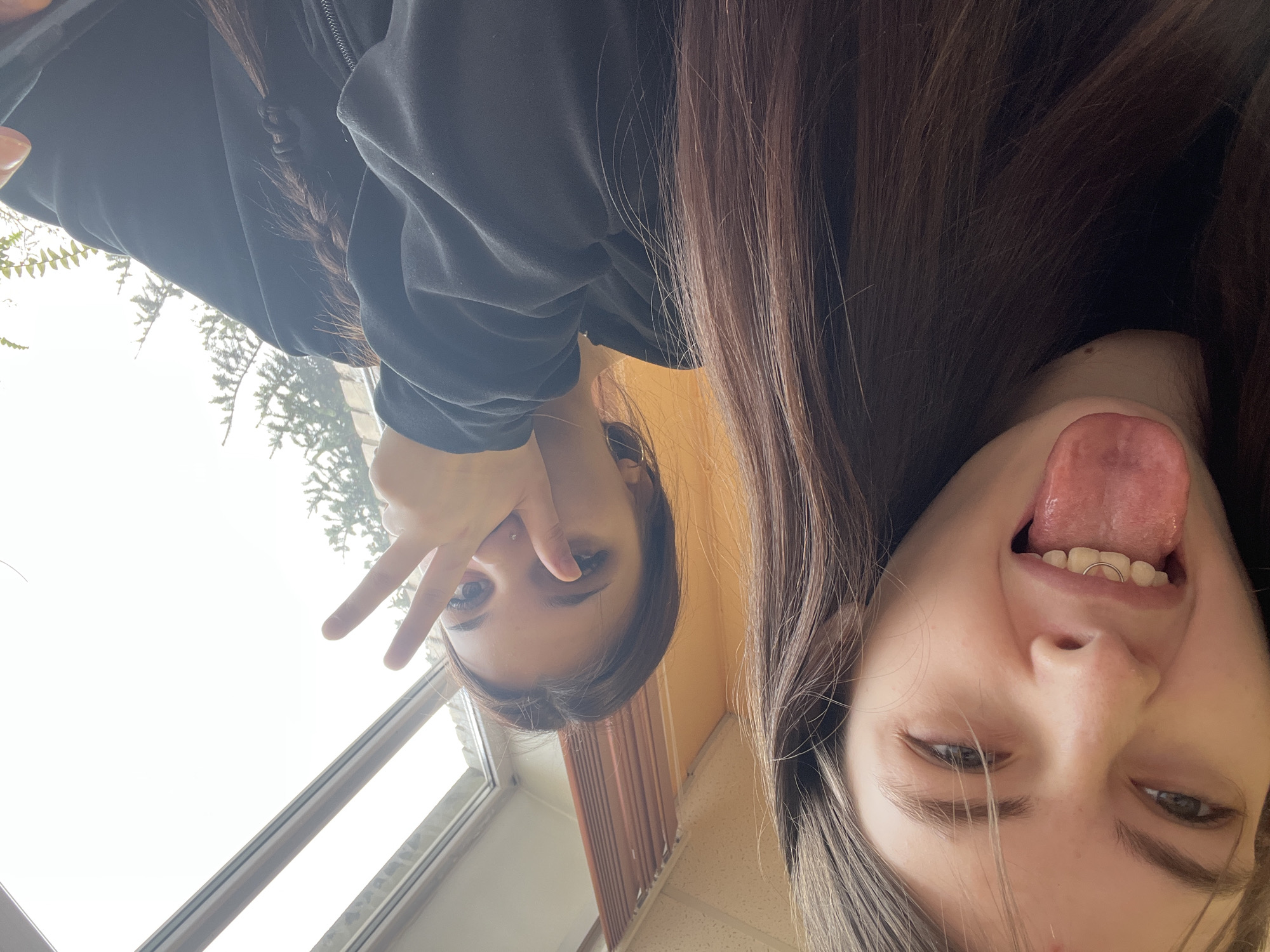 Two young women are visible in the image, likely taking a selfie. The image appears to be inverted. The woman on the left side of the image is holding up two fingers in a "peace" sign. The woman on the right has her tongue out, and is wearing a lip ring. They are indoors, near a window. It is difficult to ascertain the location as St. Petersburg, Russia without additional visual cues.