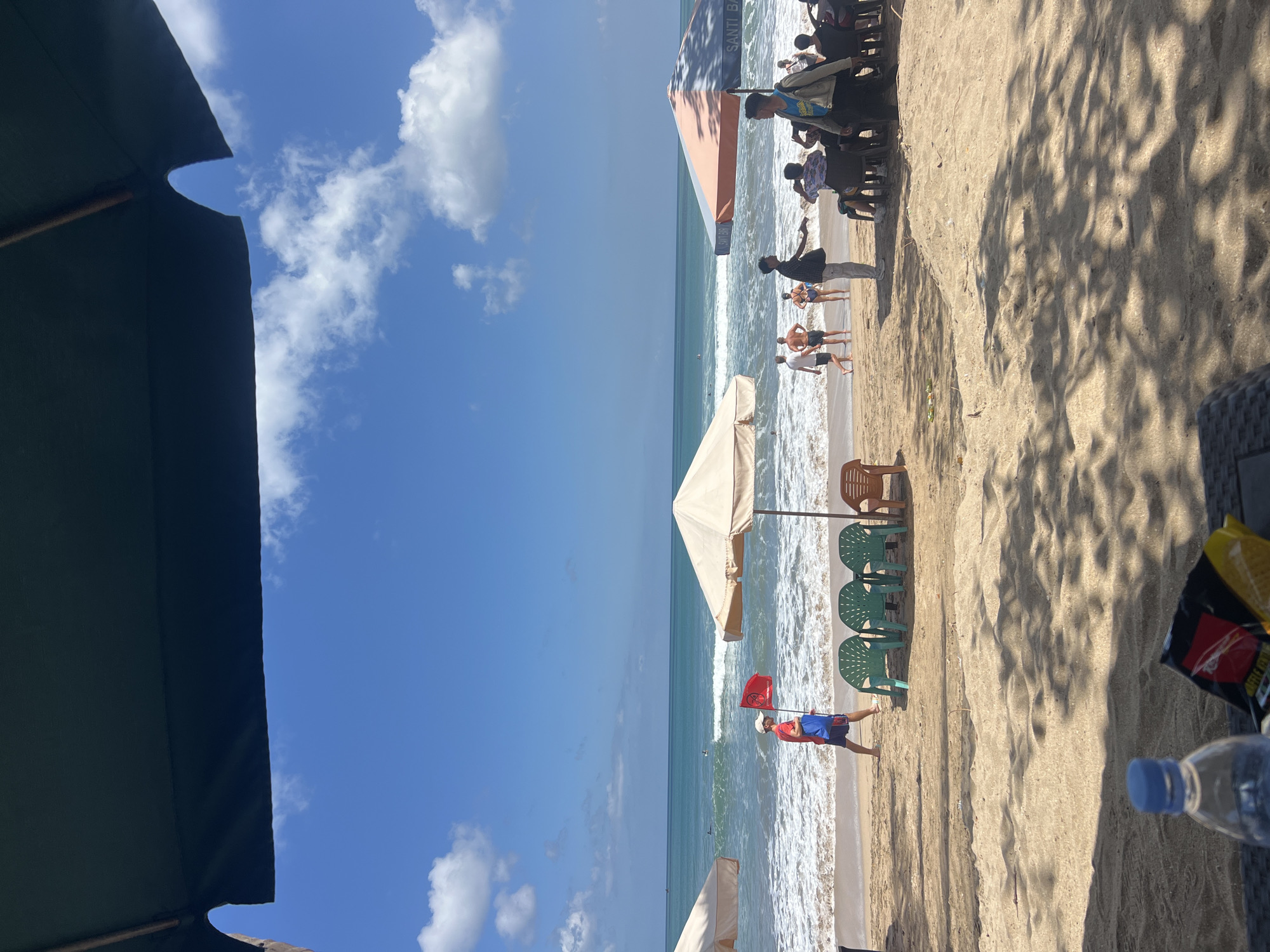 The image depicts a sunny day at Kuta Beach, Indonesia. The foreground is dominated by sandy beach with shadows cast by umbrellas. In the midground, people are seen walking and relaxing on the beach. A man in a red shirt and shorts walks with a red flag and a blue bag, possibly a lifeguard. Several beach chairs are set up on the sand. In the background, the turquoise ocean meets the shore with gentle waves, and surfers can be seen in the distance. The sky is a clear blue with scattered white clouds. Umbrellas are providing shade on the beach, with one umbrella on the right displaying "SANTI BALI". The scene captures a typical day at a popular tourist beach.