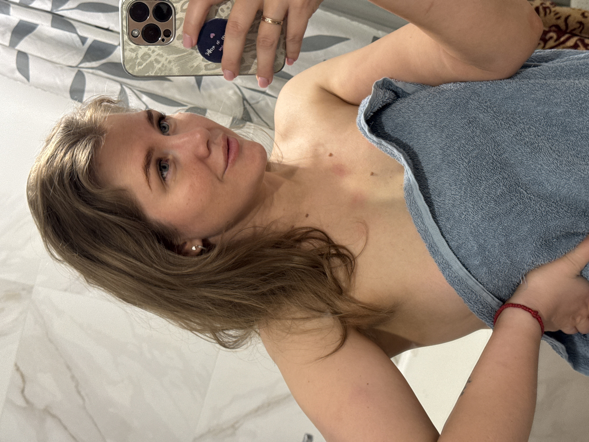 A selfie shows a fair-skinned woman wrapped in a blue towel. She is in a bathroom with marble-like walls and a gray and white patterned curtain. The woman has light brown hair and blue eyes. She is holding a smartphone with a patterned case, presumably taking the picture. A red bracelet is visible on her wrist. The setting appears to be indoors, likely a residential bathroom, possibly in the city of Ферма, Russia.