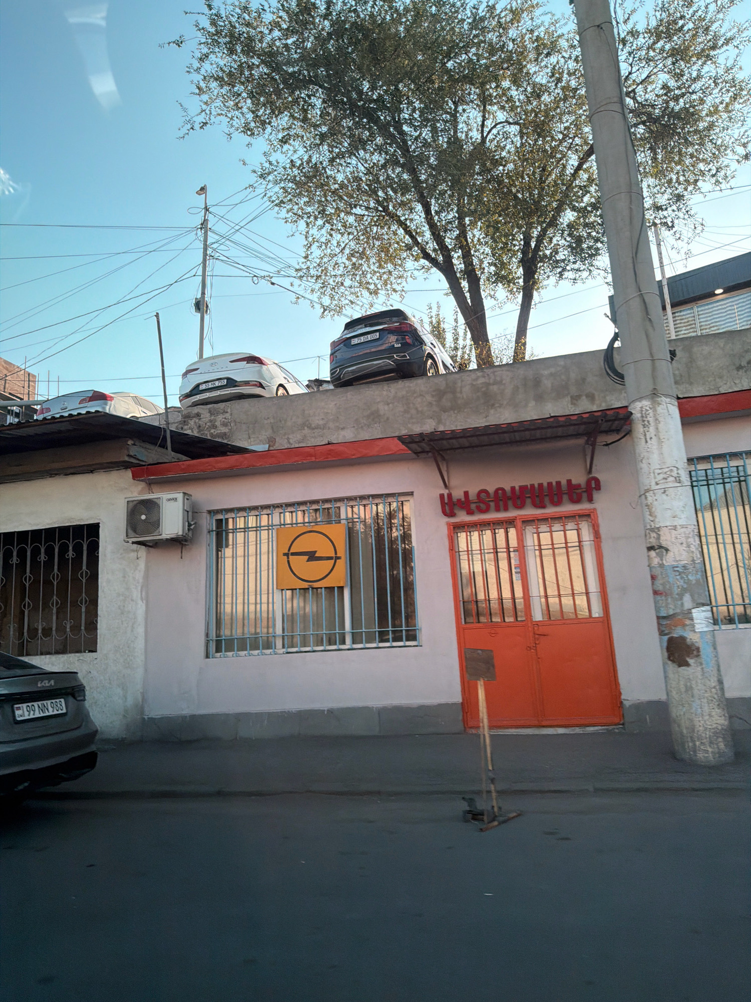 The image depicts a street scene in Yerevan, Armenia, with a building facade and cars parked on a higher level. The building has a light-colored wall with barred windows and an orange door. An air conditioning unit is mounted on the wall above one of the windows. A yellow sign with a black Opel logo is visible through a window. Above the building, several cars are parked on what appears to be a rooftop or elevated platform. These cars include a white Hyundai, a dark-colored SUV, and another car partially visible. Electrical wires and a utility pole are prominent in the sky. A dark-colored KIA is parked on the street in the foreground. The overall scene is urban and appears to be during daylight.