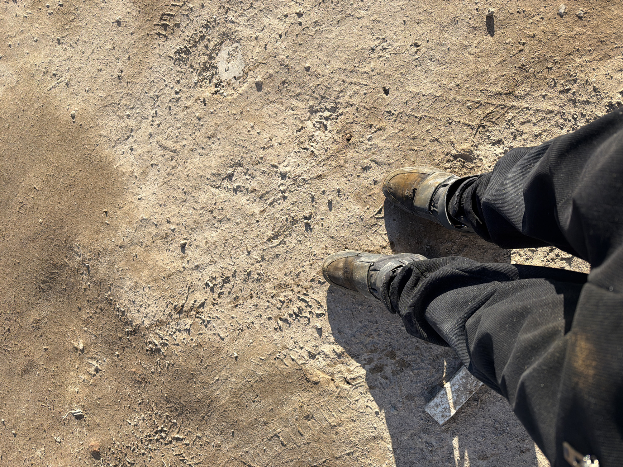 A top-down shot shows a person standing on a dirt or gravel surface. The person is wearing black corduroy pants and tan or gray shoes with straps. The surface is uneven, with patches of darker and lighter material, and appears to be marked with tire tracks and footprints. A metallic object, possibly a small tool or piece of debris, is visible near the person's right leg. The location is unspecified, and there's no mention of Chaikovsky, Russia, or any specific city.