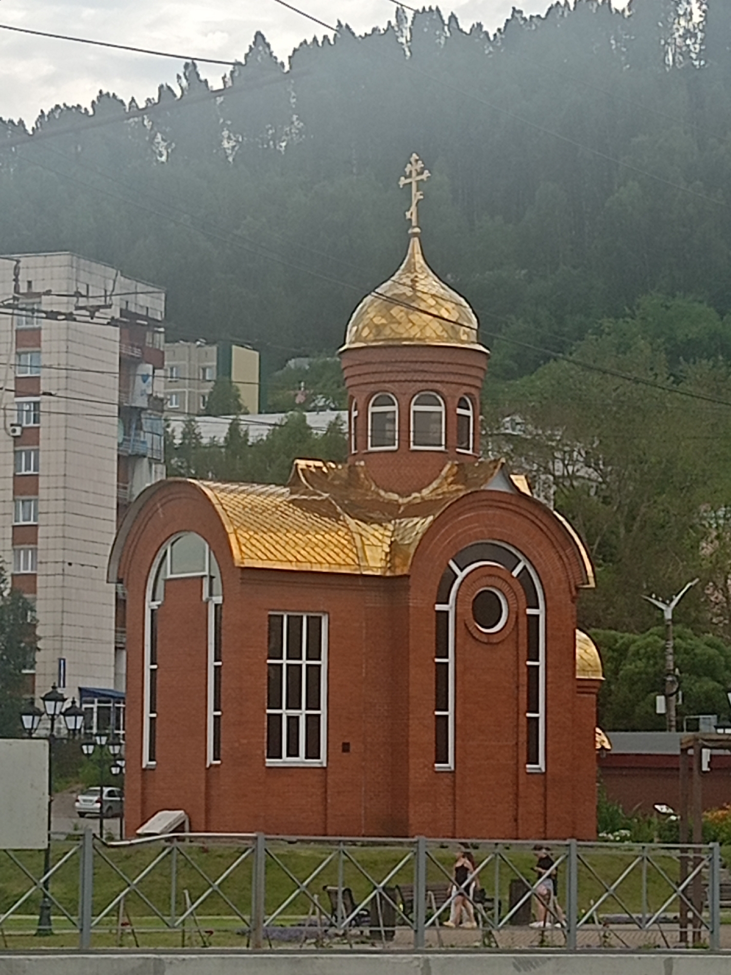 The image depicts the Church of the Archangel Michael in Zlatoust, Russia. The church is a relatively new brick structure with a golden dome topped by a cross. It features arched windows and a prominent central dome. In the background, there are several multi-story apartment buildings and a forested hill. The foreground shows a metal fence, benches, and two individuals walking. The sky is overcast.