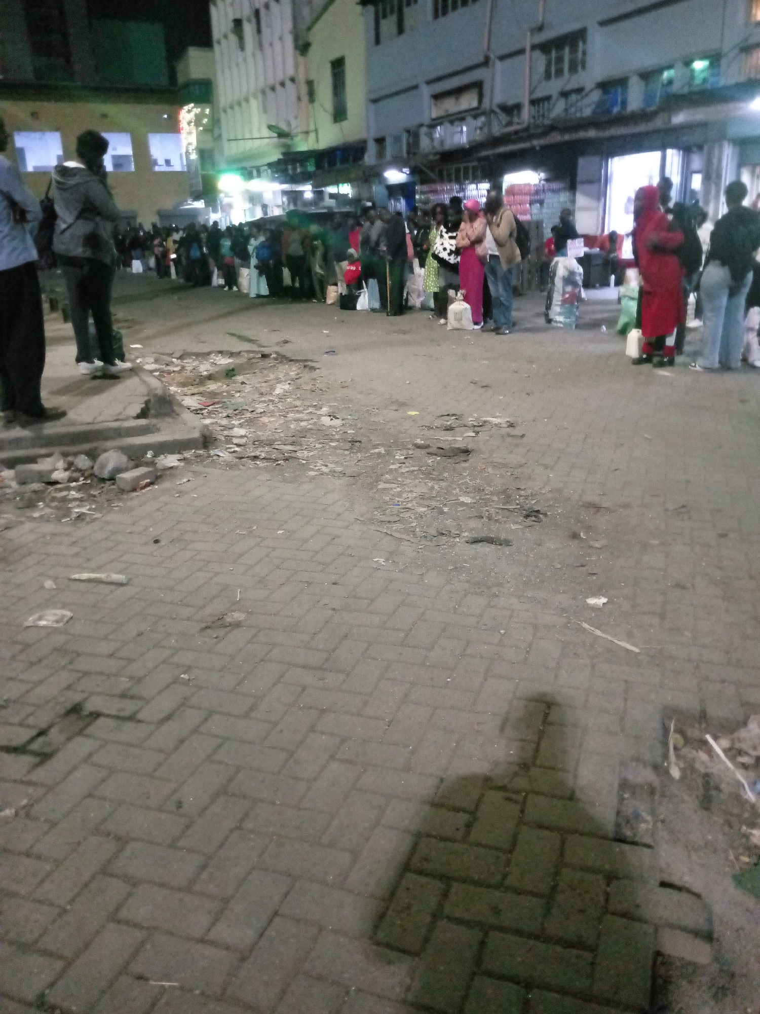 The image shows a nighttime scene in Nairobi, Kenya. A long queue of people is visible outside a building. Many people are carrying bags or other items. The street is paved with interlocking bricks and has debris scattered across it. A shadow of a person taking the photo is visible in the foreground. The building behind the queue has bright lights, suggesting a commercial establishment.