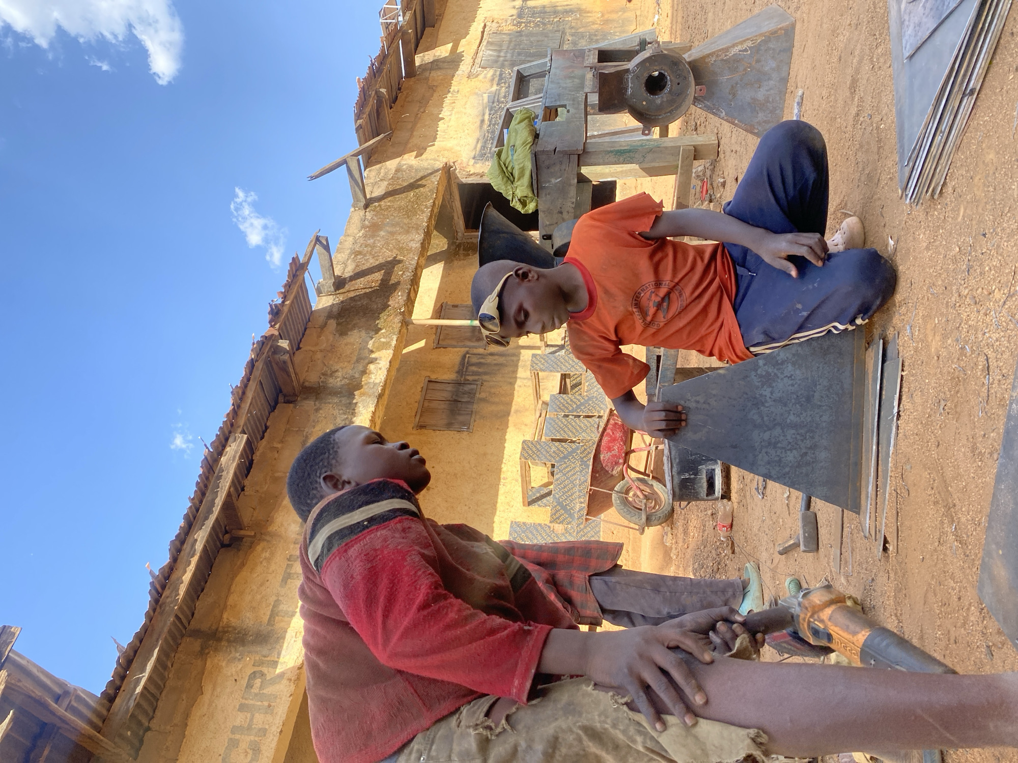 Two young men are sitting outdoors, engaged in metalwork, in Kuru Dungus, Nigeria. One wears an orange shirt and dark pants, the other a red shirt and brown pants. They are surrounded by metal sheets and tools. A building is in the background, and the ground is dirt.