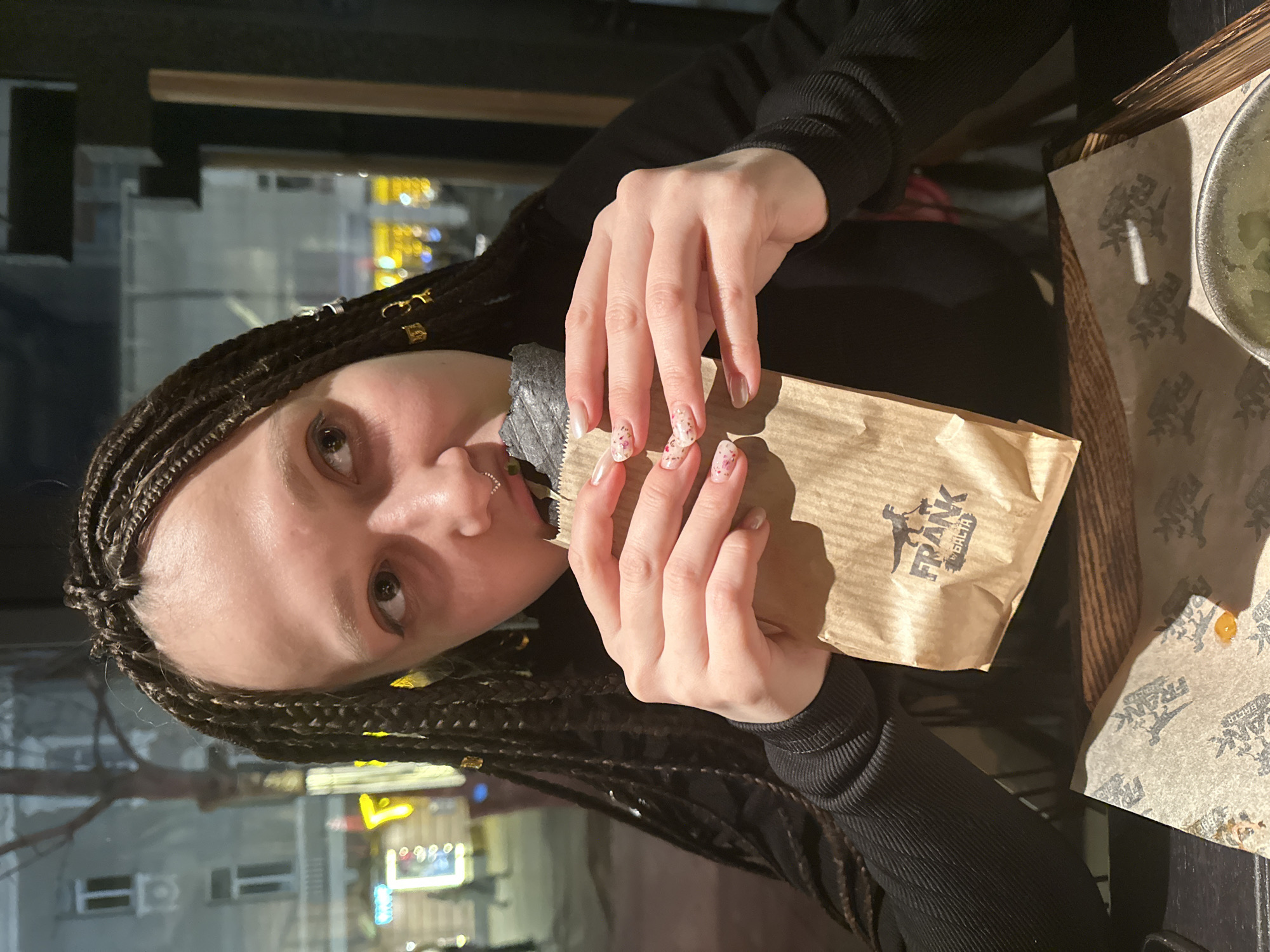 A woman with braided hair and decorative golden accessories in her braids holds a brown paper bag labeled "FRANK БЛЧТА" in her hands, with a restaurant-branded placemat visible on the table. A bowl of food sits partially visible on the right edge. The backdrop consists of a blurred urban night scene, discernible through a window, indicative of the city of Saratov, Russia, where "Frank" restaurants can be found.