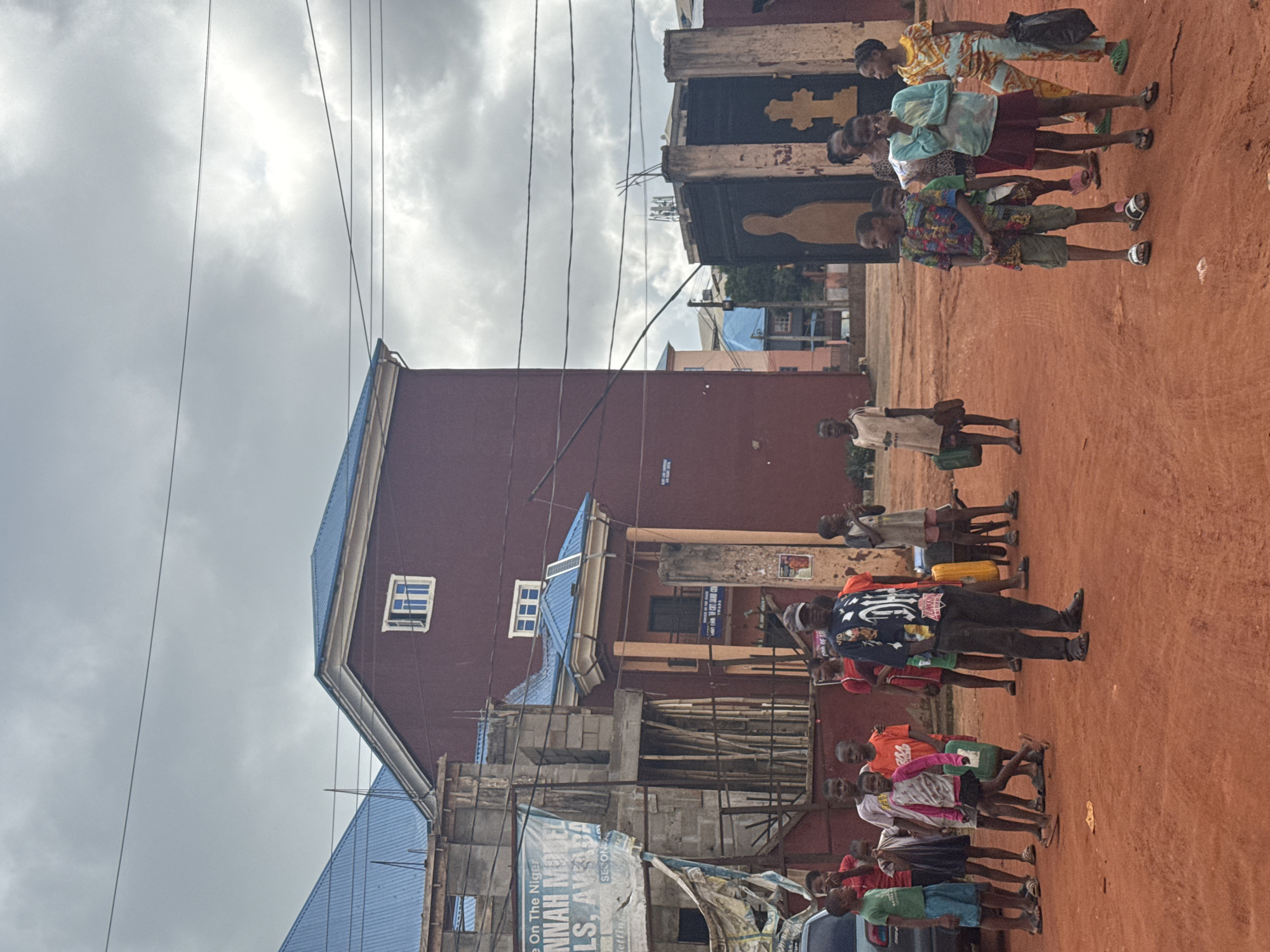 The image depicts a street scene in Onitsha, Nigeria, under an overcast sky. A group of children, some carrying green containers, are gathered on a reddish-brown dirt road in front of buildings. One building, partially under construction, has a sign that reads "HANNAH MOTEL & LODGING, ALS, AVADA." Adjacent to this, a larger maroon building with a blue roof has arched walkways and ornate entrances. To the right, a black metal gate with a cross motif is visible, with a silhouette of a person on it. The children are dressed in casual, colorful clothing. One adult, wearing a dark baseball cap and a patterned shirt, stands in the foreground, facing away from the camera. The overall impression is one of everyday life in a developing urban environment.