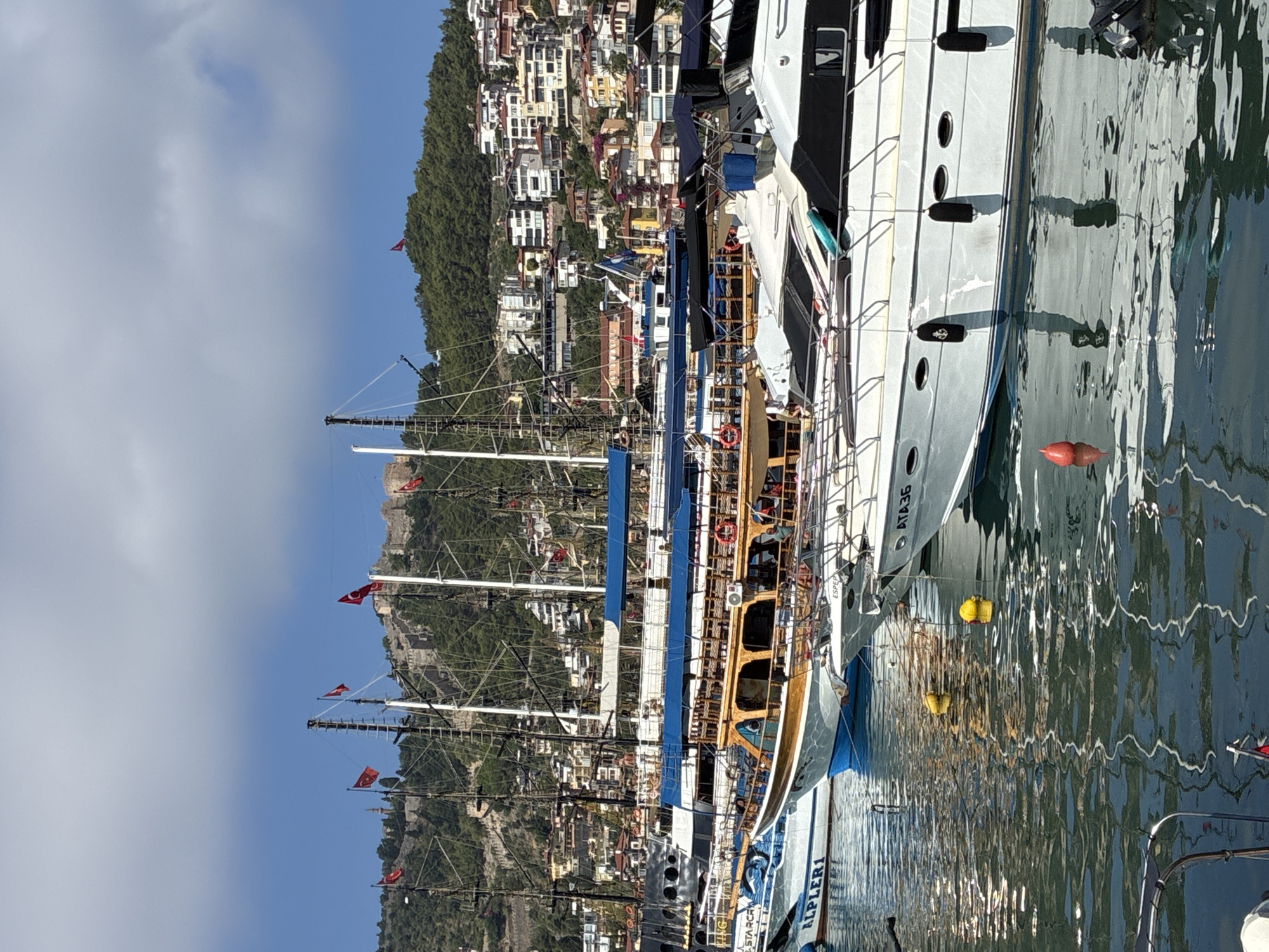 The image depicts a picturesque harbor scene in Alanya, Turkey, with a variety of boats docked under a clear blue sky with scattered clouds. In the foreground, several white yachts with black accents are moored, their reflections shimmering on the water's surface. One yacht prominently displays the registration number "ATA36". A traditional wooden boat with a blue canopy is visible behind the yachts, its polished wood gleaming. Further in the background, larger sailing vessels with multiple masts stand tall, their rigging complex against the sky. The cityscape of Alanya rises up the hillside behind the harbor, characterized by densely packed buildings with red-tiled roofs, interspersed with lush green trees. Dominating the skyline is a historic castle fortress, perched high on the hill, a testament to the city's rich past. Turkish flags are visible flying from several of the boats and from the castle. The water is calm, with yellow and red buoys marking the expanse. The overall atmosphere is one of a vibrant, sun-drenched Mediterranean port.