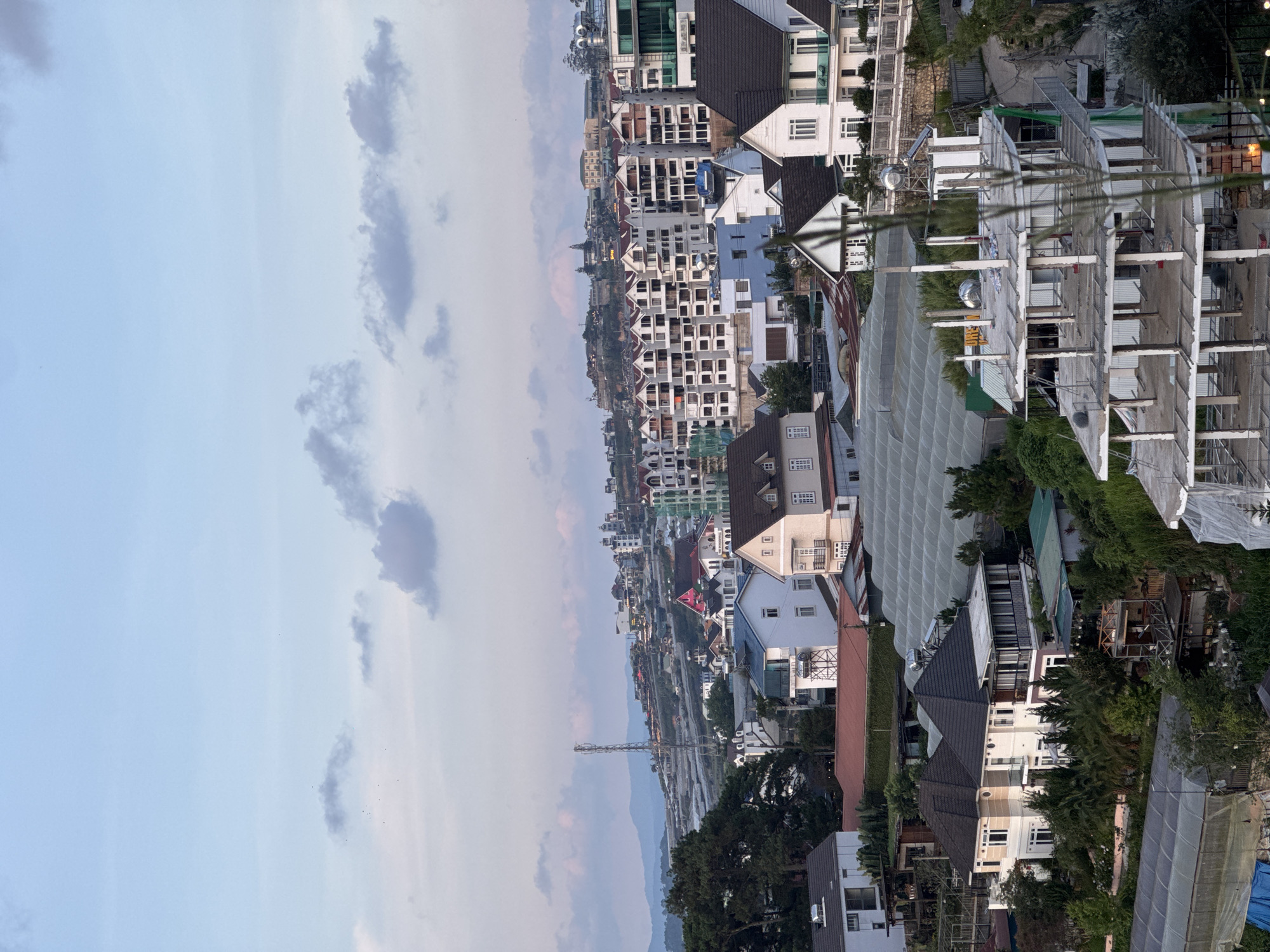 This image depicts a densely developed, hilly urban landscape in Xuân Trường - Đà Lạt, Vietnam, observed under soft, diffused light characteristic of either dawn or dusk. The sky, occupying the upper third of the frame, transitions from a pale blue at the zenith to a faint pinkish-orange hue near the horizon, dotted with scattered grey and dark grey clouds. Small dark specks, likely birds, are visible in the upper left sky.
The background features distant, hazy blue-grey mountains. Closer hillsides are extensively covered with large, semi-transparent greenhouses, indicating agricultural activity. A tall, slender cellular or telecommunications tower stands prominently on a ridge to the left of the image's vertical center. Further residential buildings are visible on these distant slopes.
The midground is dominated by a dense cluster of multi-story buildings and houses cascading down the hillsides. Many structures are white or light-colored with dark, often multi-peaked, pitched roofs. A significant portion of the central and right midground shows a complex of multi-story apartment-style buildings with repetitive window patterns and light-colored facades. Individual houses with dark roofs and light walls are interspersed, many featuring balconies and multiple windows. Numerous greenhouses with arched or corrugated roofs are integrated into the urban fabric. Several rooftops display metallic water tanks and solar water heating panels.
In the foreground, more residential properties are visible, including two prominent, large, light-colored houses with dark, complex roofs on the left. On the lower right, the exposed concrete skeleton of a multi-story building under construction is a prominent feature, revealing pillars, floor slabs, and visible rebar. Some scaffolding or support structures are discernible around this incomplete building. Sparse, out-of-focus green foliage partially obscures the extreme lower right and edges of the foreground. Throughout the scene, various trees and bushes provide greenery, often surrounding houses or growing on terraced sections of the hills. No people or discernible active interactions are visible in the image.