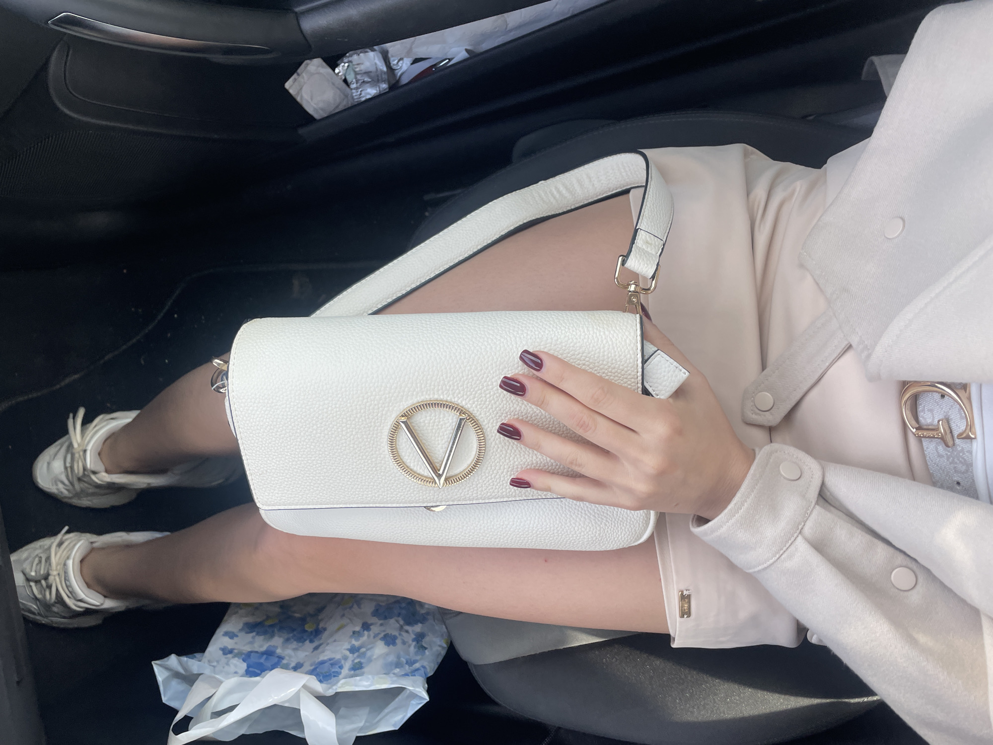 A high-angle shot captures a person seated in the passenger seat of a car, likely in Sofia, Bulgaria. The perspective focuses on their lap, legs, and one hand holding a handbag.
The subject is wearing a light beige or cream-colored top or dress, visible under an off-white jacket or shacket. A small metallic tag with the brand "GUESS" is visible on the cuff of the inner garment. A white belt with a gold-colored "G" buckle (also likely Guess) is fastened around their waist. Both feet are clad in white athletic sneakers with visible laces.
The person's right hand, adorned with dark red nail polish, rests on a white, textured handbag. The handbag features a prominent gold-colored "V" emblem within a circular ring on its front flap, suggesting a designer brand. A white strap with dark trim extends from the bag.
On the dark floor of the car, beneath the person's legs, there is a clear plastic bag with a blue floral pattern. In the car door's storage compartment, a crumpled silver foil wrapper and a distinct condom wrapper are visible, with partial text reading "DUREX" and "FEEL THIN." The car interior features dark seats and door panels. The scene appears to be illuminated by natural daylight, suggesting it is daytime. The person seems to be traveling or waiting inside the vehicle.