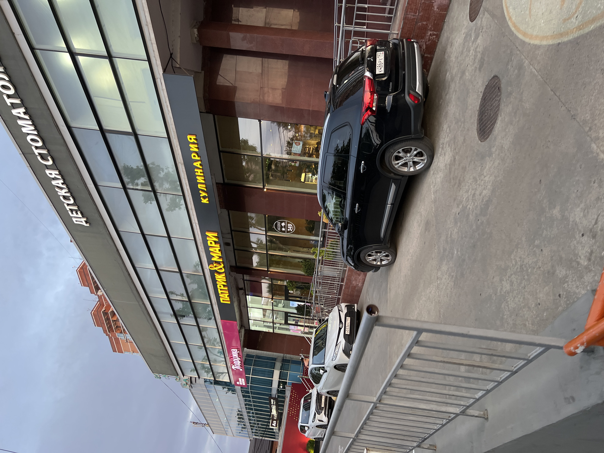 The image shows a street-level view of a modern building with large glass windows, housing several businesses. The building features a prominent sign at the top in white lettering that reads "ДЕТСКАЯ СТОМАТОЛОГИЯ" (Children's Dentistry). Below this, a dark gray facade strip displays two illuminated signs: "ПАТРИК & МАРИ" (Patrik & Mari) in yellow lettering and "КУЛИНАРИЯ" (Culinary) in yellow lettering. To the left of these signs, another pink sign reads "Подарочка" (Gift). The building's lower section is clad in dark red marble.
In front of the building, there are several vehicles. A black SUV is parked to the right, with its rear visible. To the left, two white cars are parked, a sedan in the foreground and another car behind it. The scene appears to be in a commercial area, with a paved ground surface and metal railings in the foreground. The sky is overcast, suggesting either early morning or late afternoon. Based on the signage in Russian and the architectural style, the location is likely a city in Russia, and given the context provided to similar images, it can be identified as Krasnodar, Russia.