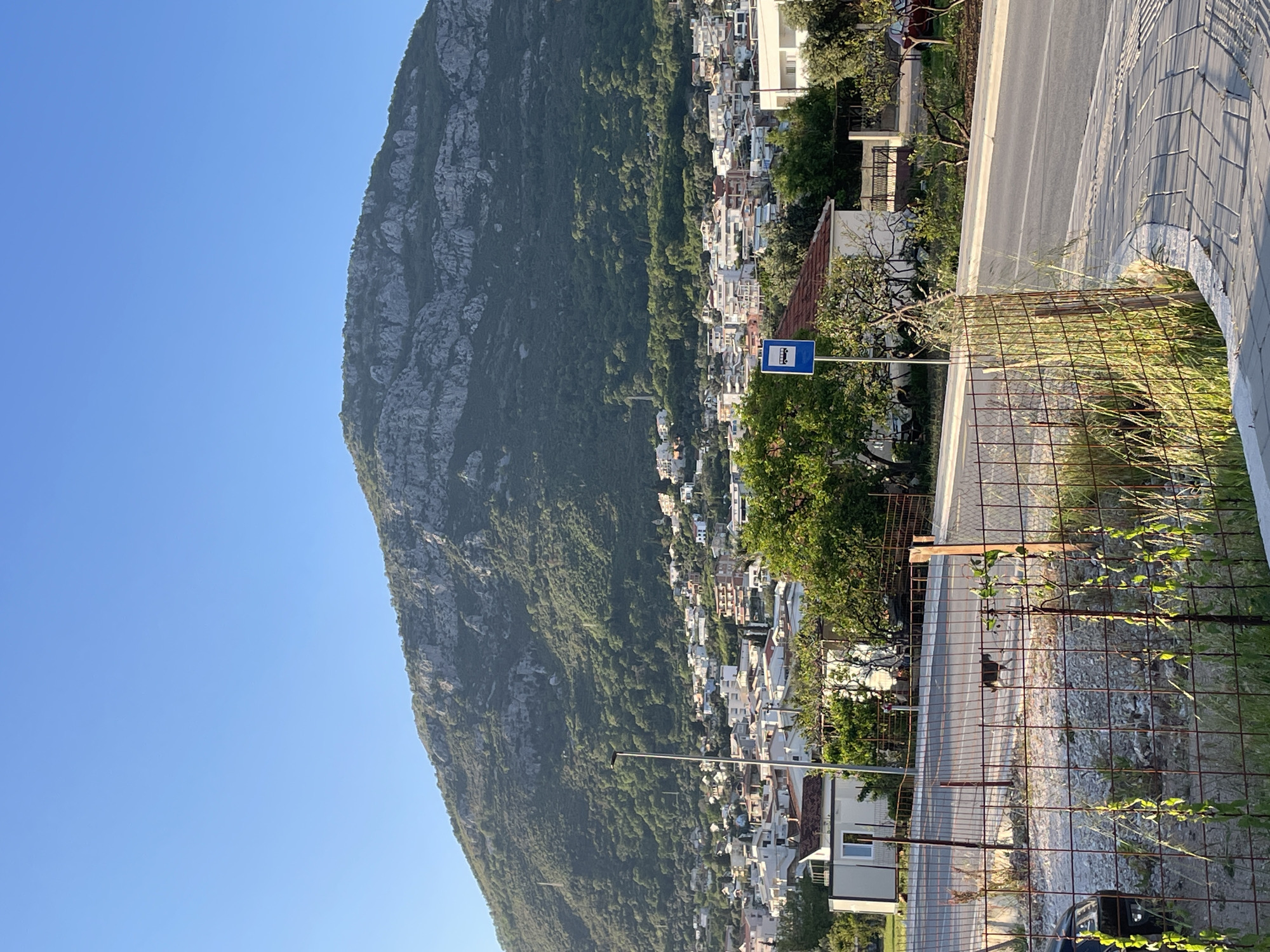 This landscape photograph captures a vibrant mountain and coastal town scene under a clear, bright blue sky. A prominent, rugged mountain, densely covered in green trees and foliage with visible grey rock faces, dominates the left side of the frame, its steep slopes extending towards the right.
Along the base and lower slopes of this mountain, a town is spread out, characterized by numerous white and light-colored buildings, most appearing to be residential, with some featuring terracotta-colored roofs. The structures are tightly packed, indicating a well-established settlement.
In the mid-ground and foreground on the right, a paved road with a light-colored curb is visible, running somewhat parallel to the mountain. A grey tiled sidewalk or paved area borders the road on the far right. A rusty metal mesh fence spans horizontally across the lower foreground, with green grass and small plants growing through its mesh. A small, dark-colored car is partially visible at the very bottom right, behind the fence.
A notable object in the mid-right is a blue rectangular sign with a white bus icon, mounted on a pole, indicating a bus stop. The entire scene is brightly illuminated by natural sunlight, suggesting a clear day, likely morning or late afternoon, given the visible shadows. The serene setting is located in Bar, Montenegro. No people are visible.