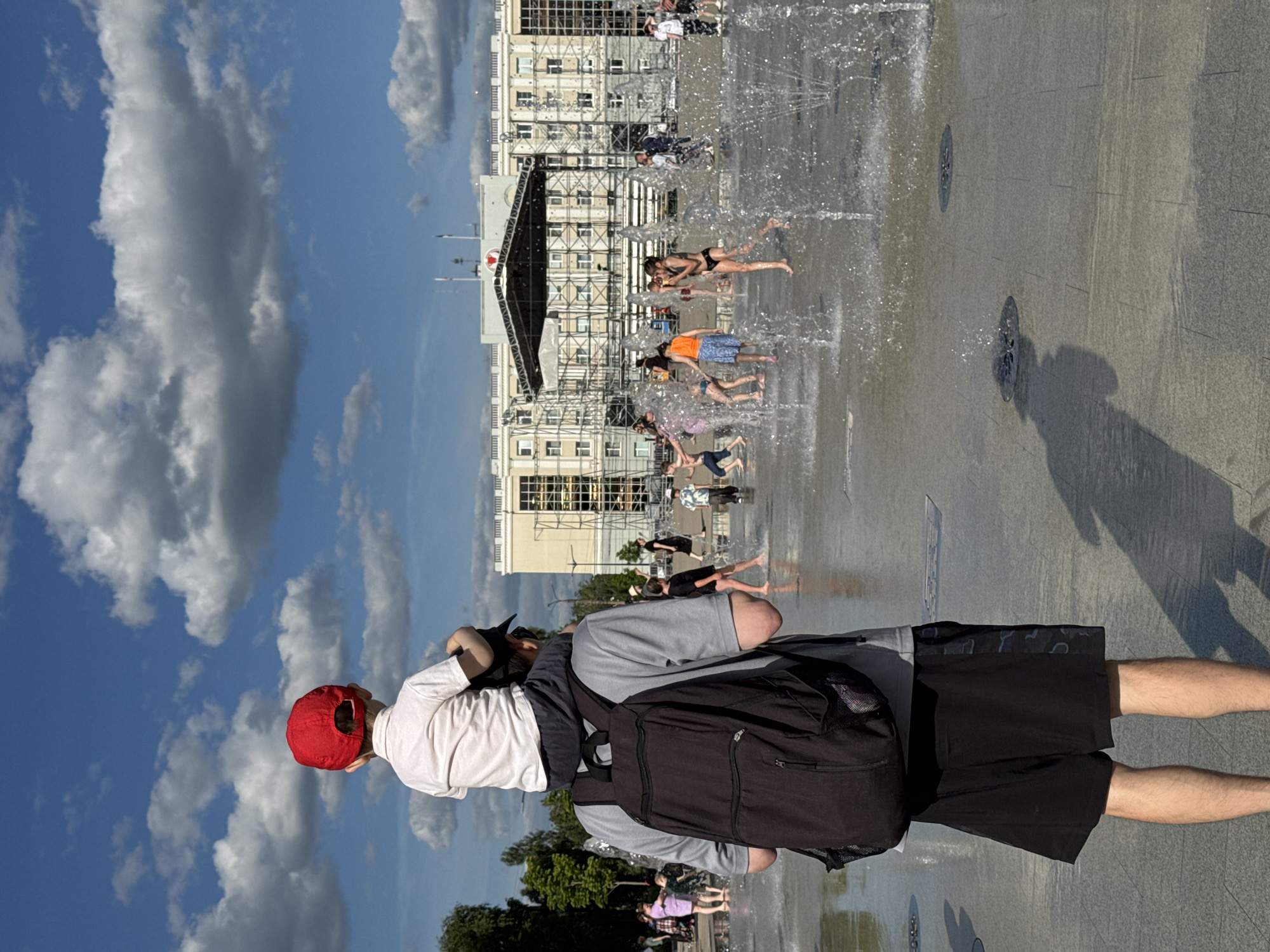 The image depicts a public square in Izhevsk, Russia, specifically in front of the Government House of Udmurtia, featuring a flat-level water fountain under a clear blue sky with scattered cumulus clouds. In the immediate foreground, a male adult, viewed from behind, wears a grey t-shirt, black shorts with a subtle dark camouflage pattern on the side, and a black backpack. He carries a young child on his shoulders. The child, also seen from behind, wears a white t-shirt and a red baseball cap, looking towards the middle ground.
The middle ground is occupied by a walk-through fountain with multiple water jets spraying upwards from the paved ground. Numerous individuals, predominantly children, are actively playing in the splashing water. Visible figures include a girl in a black two-piece swimsuit and another person in orange shorts and top, both interacting with the water. The ground in this area is wet and reflective.
The background features the large, multi-story, light-colored Government House of Udmurtia. In front of this building, a large temporary event stage with a dark canopy and supporting scaffolding is erected. A red heart symbol is mounted above the stage. Flags, including what appears to be the Russian national flag and another flag with vertical stripes (consistent with the flag of Udmurtia), are visible on poles atop the Government House. Additional scaffolding is present around parts of the building. Green trees and foliage are visible on the left side of the frame. Long shadows of people are cast on the wet ground, indicating bright sunlight.