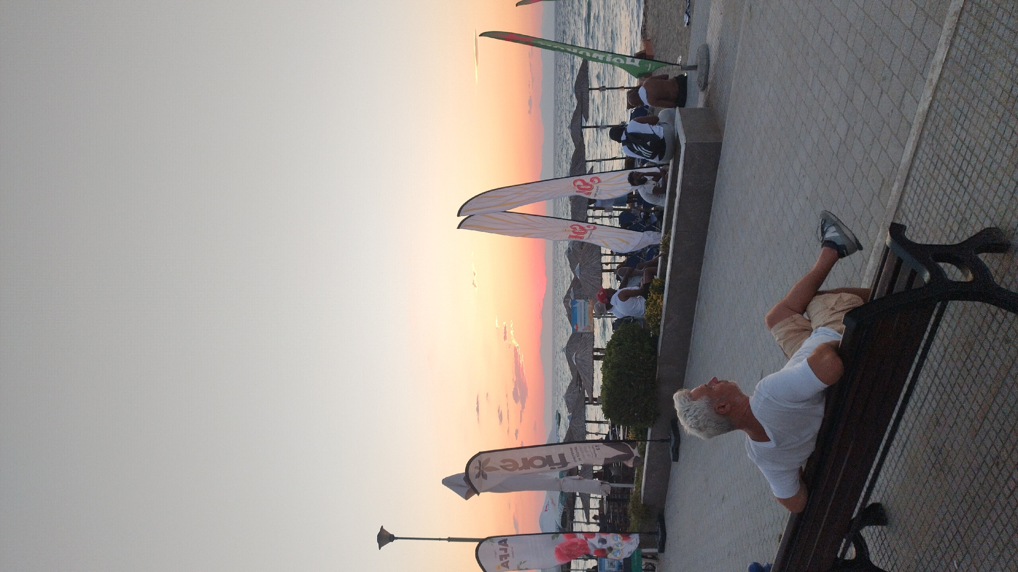 The image depicts a sunset scene on a beach in Perea, Greece. In the foreground, a man with gray hair is seated on a dark wooden bench, facing the sea and wearing a white t-shirt and tan shorts. The background shows a beach with straw umbrellas, people sitting on lounge chairs, and various flags. The sky is a gradient of pale orange and pink, indicative of sunset, with distant mountains visible on the horizon. The paved walkway in front of the bench is laid with grey square tiles.