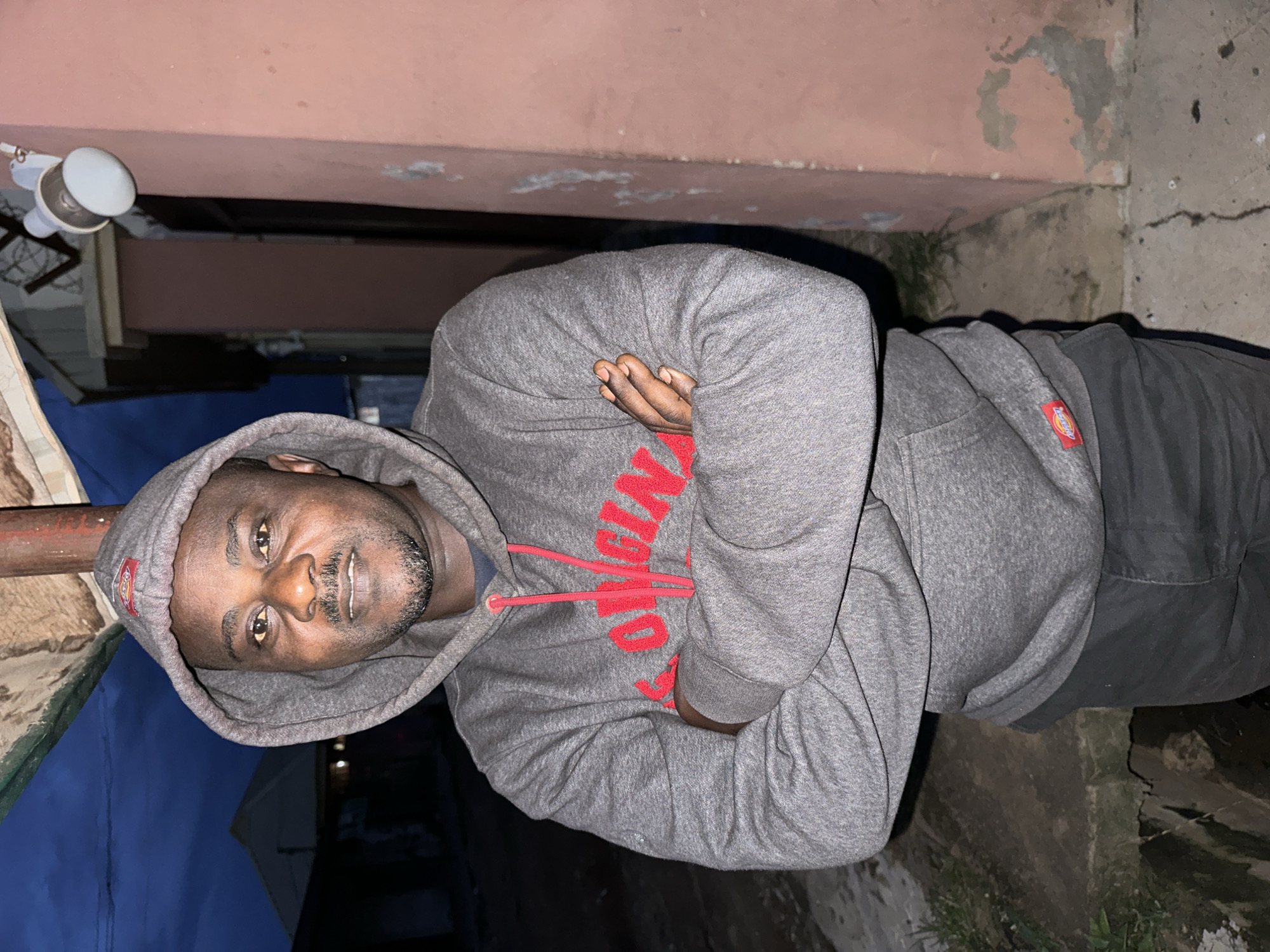 A medium shot captures a Black man with dark skin, a beard, and a mustache, looking directly at the camera. He is wearing a gray hooded sweatshirt with red lettering that reads "ORIGINAL" and dark gray pants. His arms are crossed, and he stands outdoors in front of a coral-colored wall, which appears to be part of a building. Behind him, a dark blue sky is visible with a white outdoor light fixture mounted on the wall. The ground in front of him is concrete with some patchy grass. The scene is lit by artificial light, suggesting it is either dusk or nighttime. The man's posture is relaxed, and his expression is neutral. The overall setting appears to be an urban environment, potentially residential, given the building and outdoor lighting. While the provided information mentions Narayi, Nigeria, this specific location is not visually discernible from the image content alone.