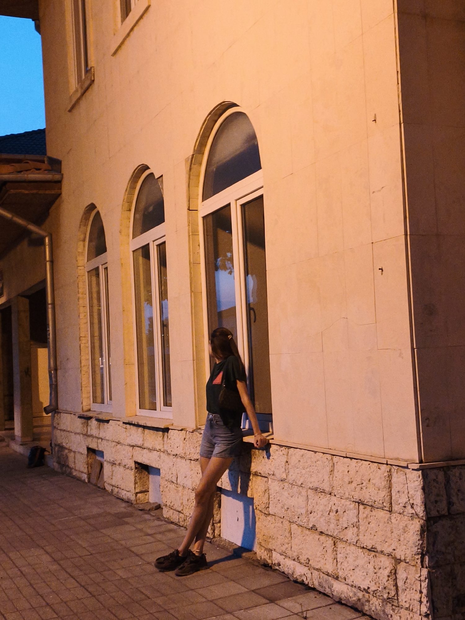 A young woman with dark hair, wearing a dark t-shirt and denim shorts, stands leaning against a light-colored stone building with arched windows. The building is illuminated by warm artificial light, casting shadows on the tiled sidewalk. A dark backpack is visible on the sidewalk to the left of the building. The scene appears to be in the city of Lovech, Bulgaria, likely during the evening or nighttime given the lighting and the blue sky visible in the upper left corner.
