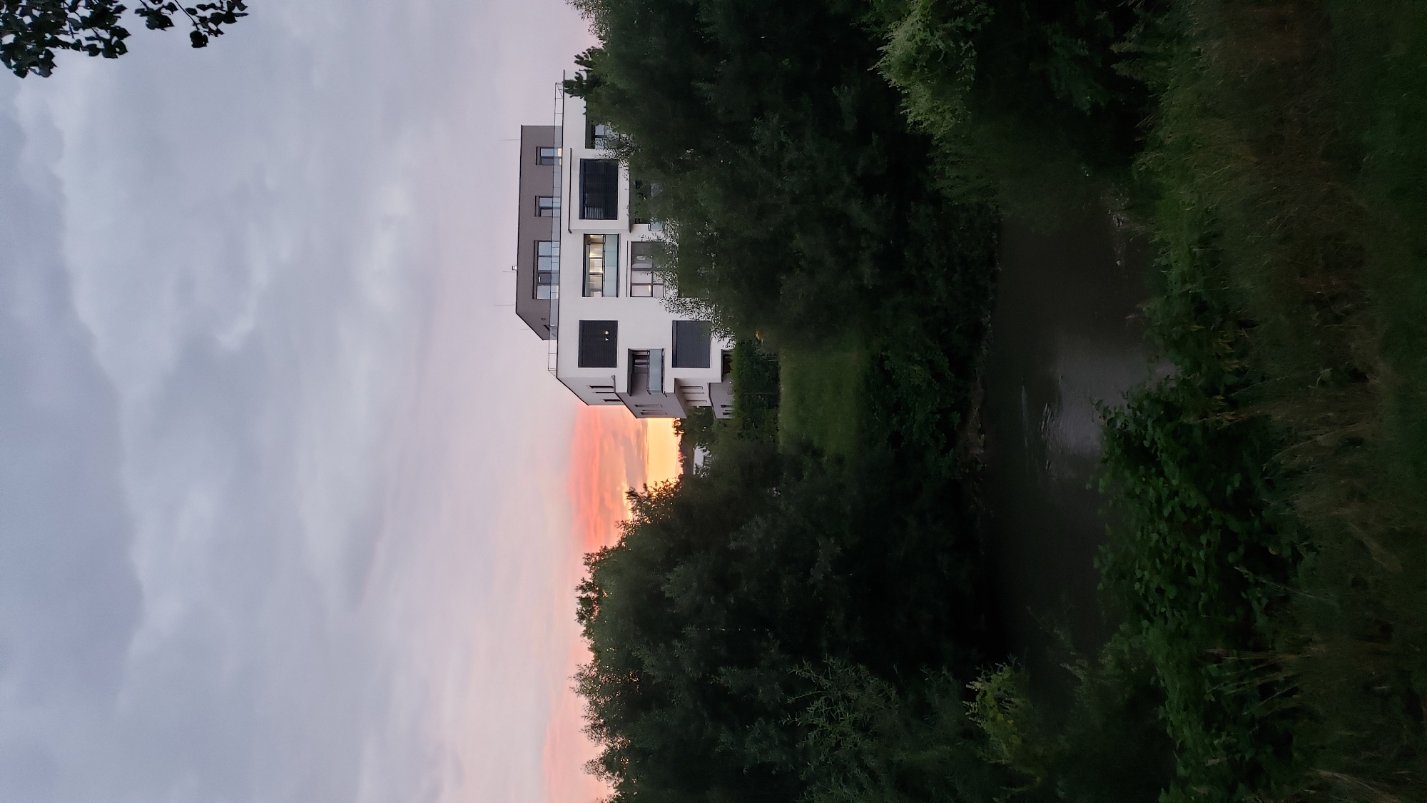 The image depicts a modern apartment building situated amidst lush greenery, with a body of water in the foreground. The sky is overcast with hints of pink and orange hues, suggesting either sunrise or sunset. The building itself is multi-story, featuring clean lines, large windows, and balconies. Dense trees and bushes surround the building and line the banks of the water. The water appears calm and dark, reflecting the sky and surrounding vegetation. The scene conveys a sense of tranquility and modern living integrated with nature. The location is identified as Prešov, Slovakia.