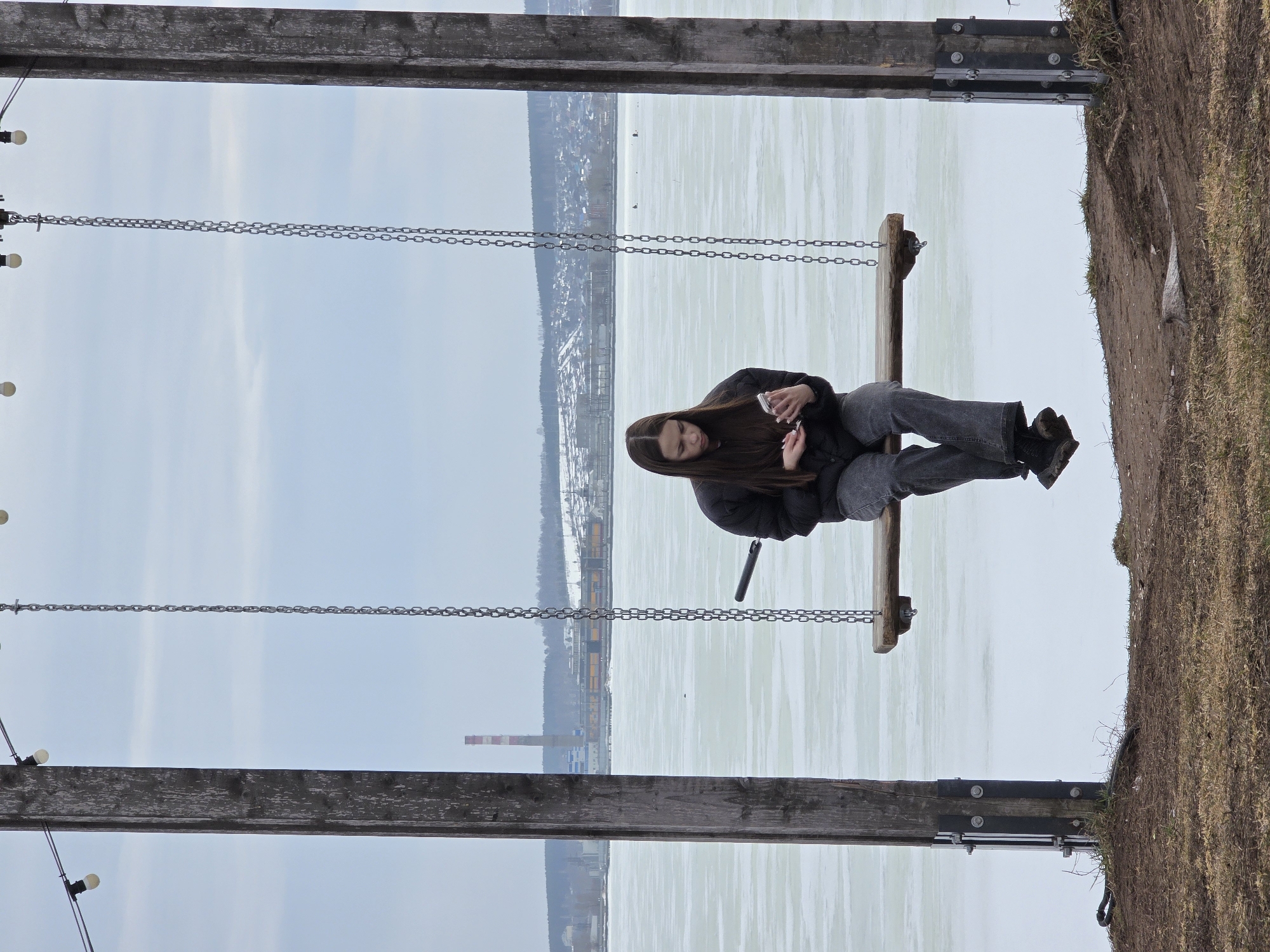 A young woman is sitting on a swing set overlooking a frozen lake in Votkinsk, Russia. She is wearing a dark jacket, grey pants, and dark boots. She is holding a phone and a vape in her hands. The swing set is made of wood and chain. The lake is covered in ice and snow. The sky is overcast. The background includes buildings and a smokestack. The ground near the swing set is covered in grass.