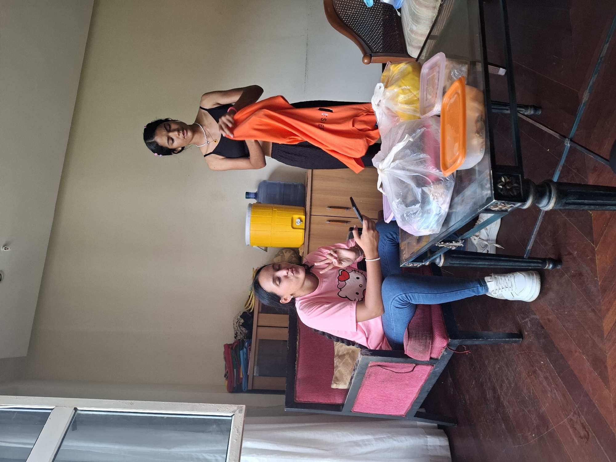 Two young women are in an indoor setting, possibly a living room or common area. The woman on the left, seated in a red armchair, wears a pink t-shirt with a Hello Kitty graphic and blue jeans. She is holding a smartphone and has her fingers raised in a peace sign, with her lips puckered. The woman on the right, standing, wears a black tank top and black pants. She is holding an orange shirt. In the background, there is a yellow water cooler, a blue water jug, and a wooden cabinet filled with folded clothes. A glass-topped coffee table in the foreground holds several plastic containers and bags of food. The flooring is dark wood. The scene suggests a casual gathering or a moment of preparation before or after an activity. The image does not contain any information to place it in the city of Pedro González, Venezuela.