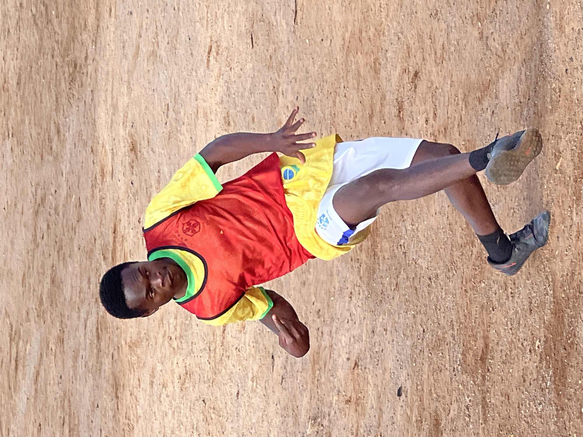 A young Black male, appearing to be in his late teens or early twenties, is captured in a dynamic pose on a dirt field, likely in Jos, Nigeria. He is wearing a yellow jersey with green trim, a red training bib with a circular emblem, yellow shorts with a blue and white design and a Brazilian flag emblem, white athletic shorts, and dark soccer cleats with orange accents. His left leg is bent at the knee and extended backward, while his right leg is forward, with the foot angled upwards, suggesting he is either dribbling a ball or in the process of taking a shot. His left arm is bent and raised, with the hand partially closed, and his right arm is extended to the side, with the fingers spread. His head is turned to the left, and his gaze is directed downwards and to the left. The field is composed of reddish-brown dirt with scattered small stones and debris. The lighting suggests it is daytime, with soft shadows indicating an overcast sky or a time of day with diffuse sunlight. The overall scene conveys a moment of athletic action and focus.