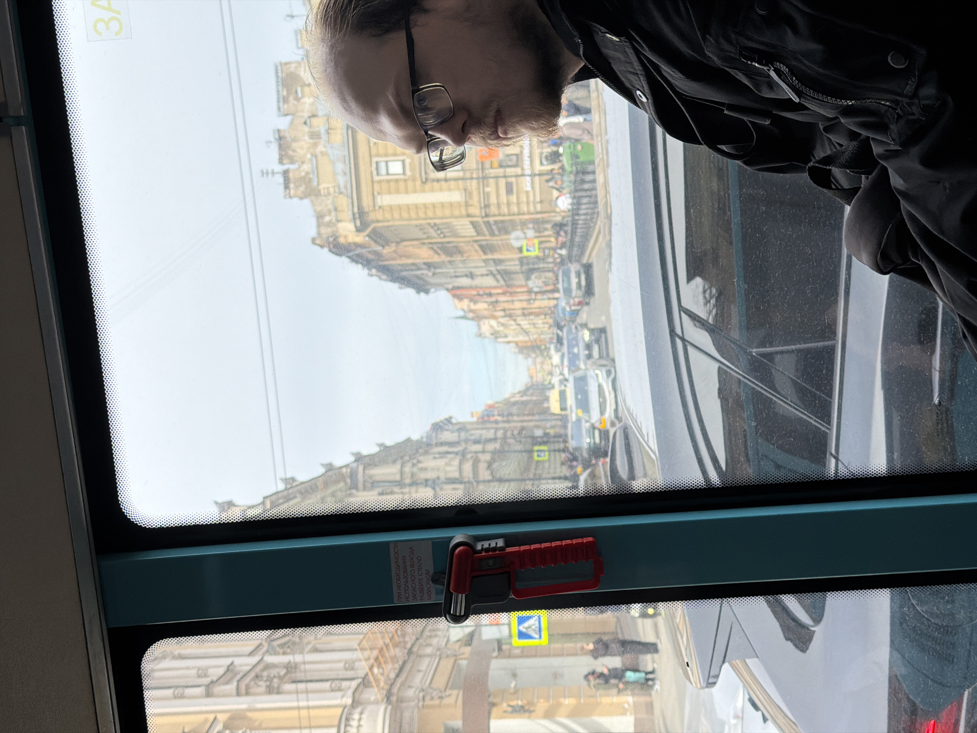 A passenger is on a tram in Saint Petersburg, Russia. The tram travels along a street lined with older buildings with ornate facades. Traffic includes cars, pedestrians, and street signs. A red emergency hammer hangs on the window frame.