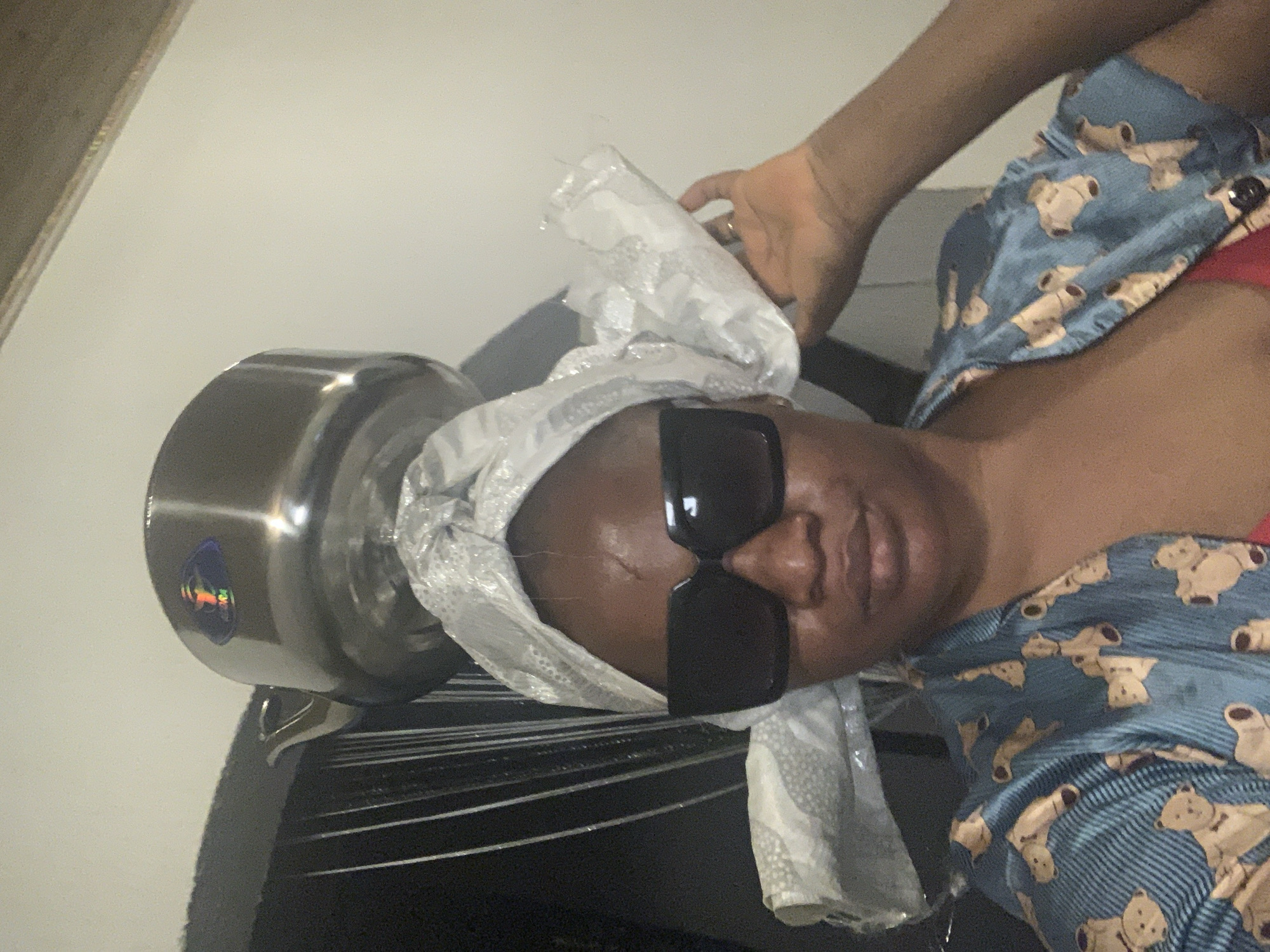 A person wearing a blue teddy bear print shirt and black sunglasses is holding a stainless steel pressure cooker in front of them. The person has a white cloth wrapped around their head. The scene appears to be indoors. No definitive information confirms the location is Barakin Akawo, Nigeria.