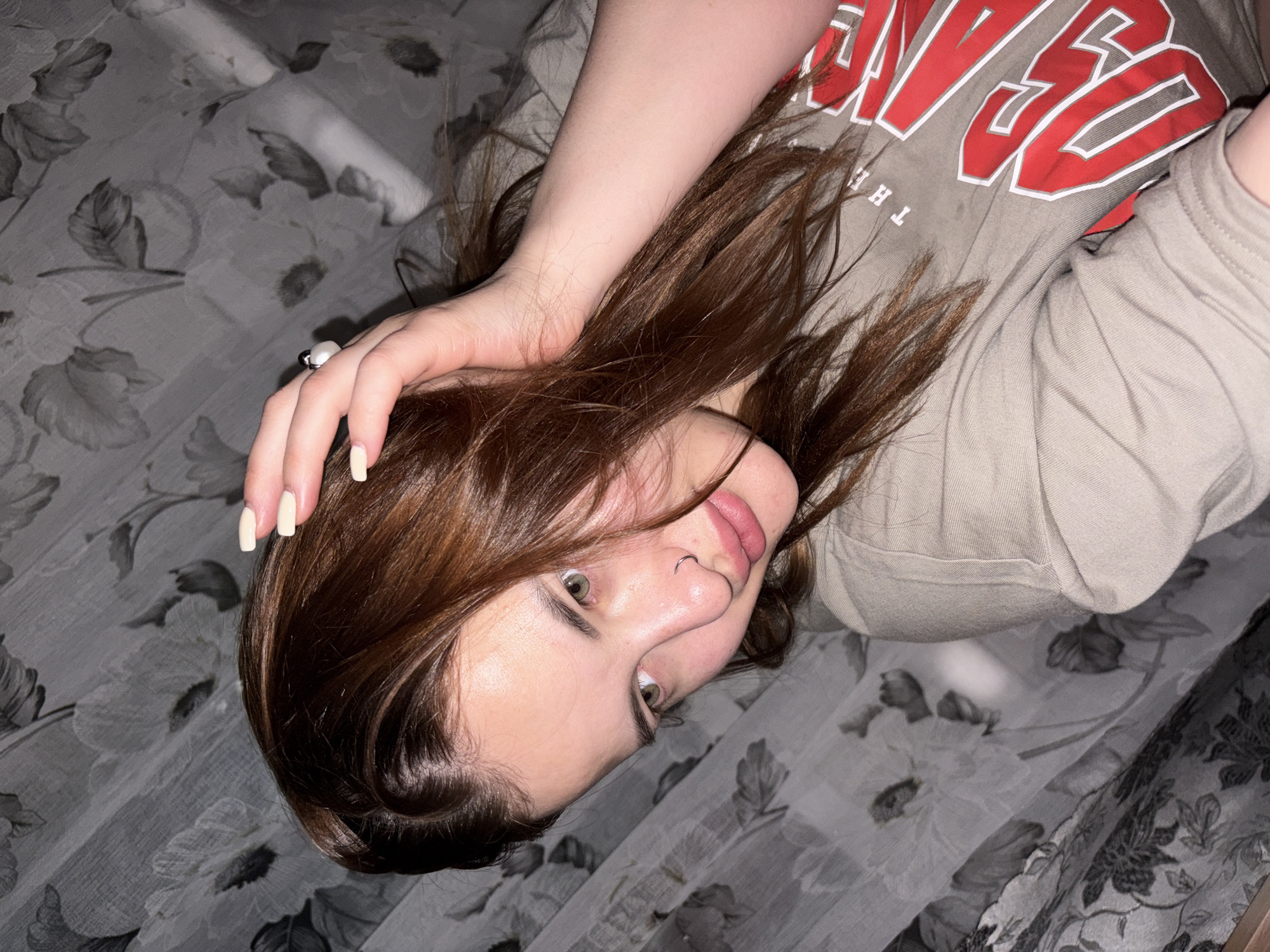 A young individual, likely female, is captured in a close-up, rotated shot, appearing to lie sideways. She has medium-length brown hair, which is slightly disheveled around her face, and striking green eyes. A metallic ring pierces her left nostril. Her lips are slightly parted. One hand, with light cream-colored manicured nails, rests on her hair near her temple. On one finger, a ring with a white and black spherical element is visible.
She is wearing a light beige t-shirt. Visible text on the shirt includes the word "THE" in white, below larger red and white letters which appear to spell out part of a word like "REDGE" or "NGE," with other partial letters forming what could be "LEAGUES" or "EVES" in a bold, collegiate-style font.
The background consists of a patterned fabric, likely a bedsheet or duvet cover, featuring a floral design in shades of grey and white. The lighting is soft, suggesting an indoor setting, possibly during the evening or in low light. The subject's expression is neutral, looking directly at the camera. The overall scene suggests a casual selfie taken in a personal space. The location context is Port-Katon, Russia.