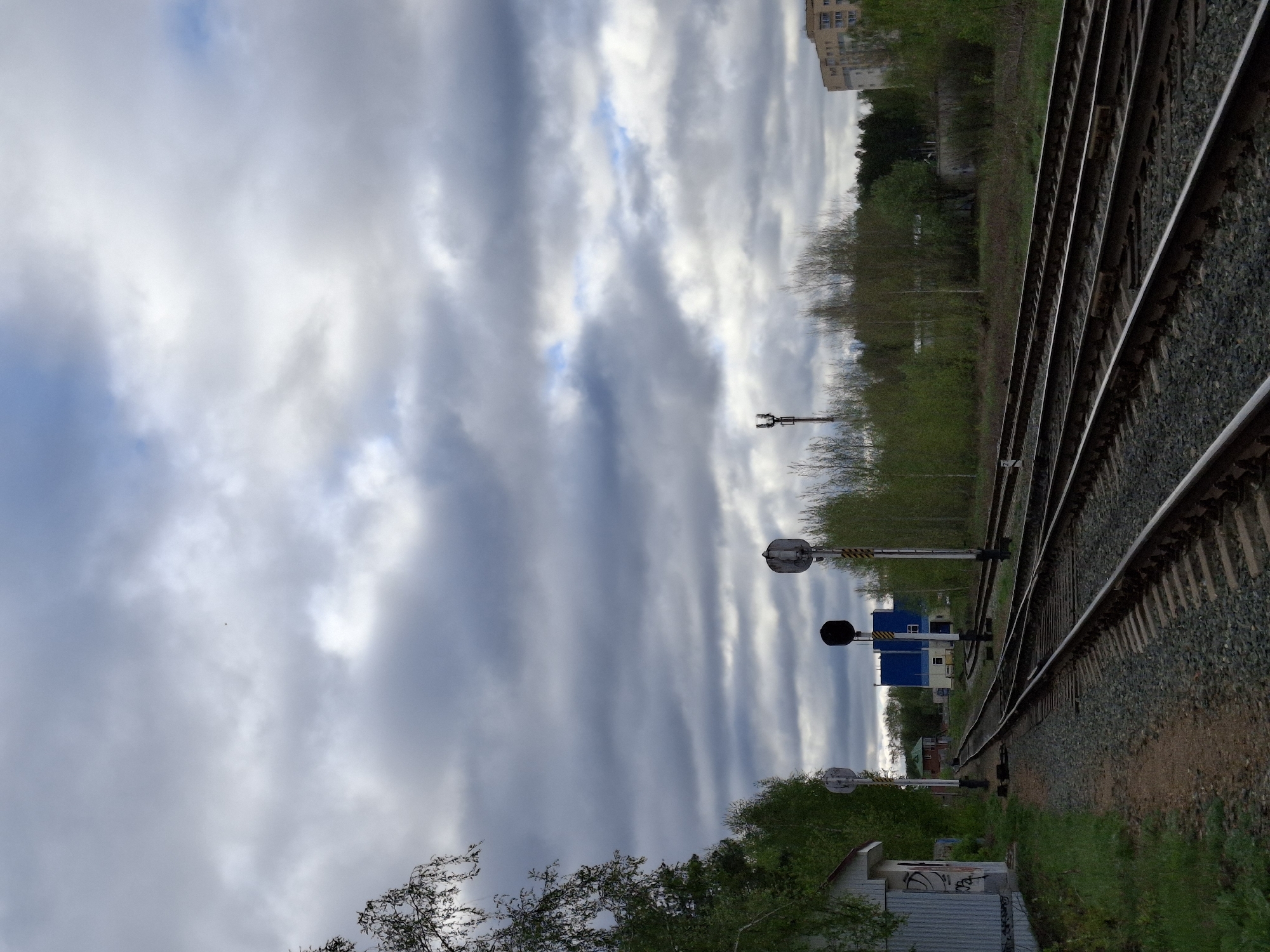 The image depicts a railway track in the city of Chaikovsky, Russia, under an overcast sky. The tracks are running along the side of the road. There are railway signals present along the railway track, including light and dark indicators. The rails appear to be made of steel. The area around the rails consists of gravel, dirt and vegetation. Buildings are visible in the background.