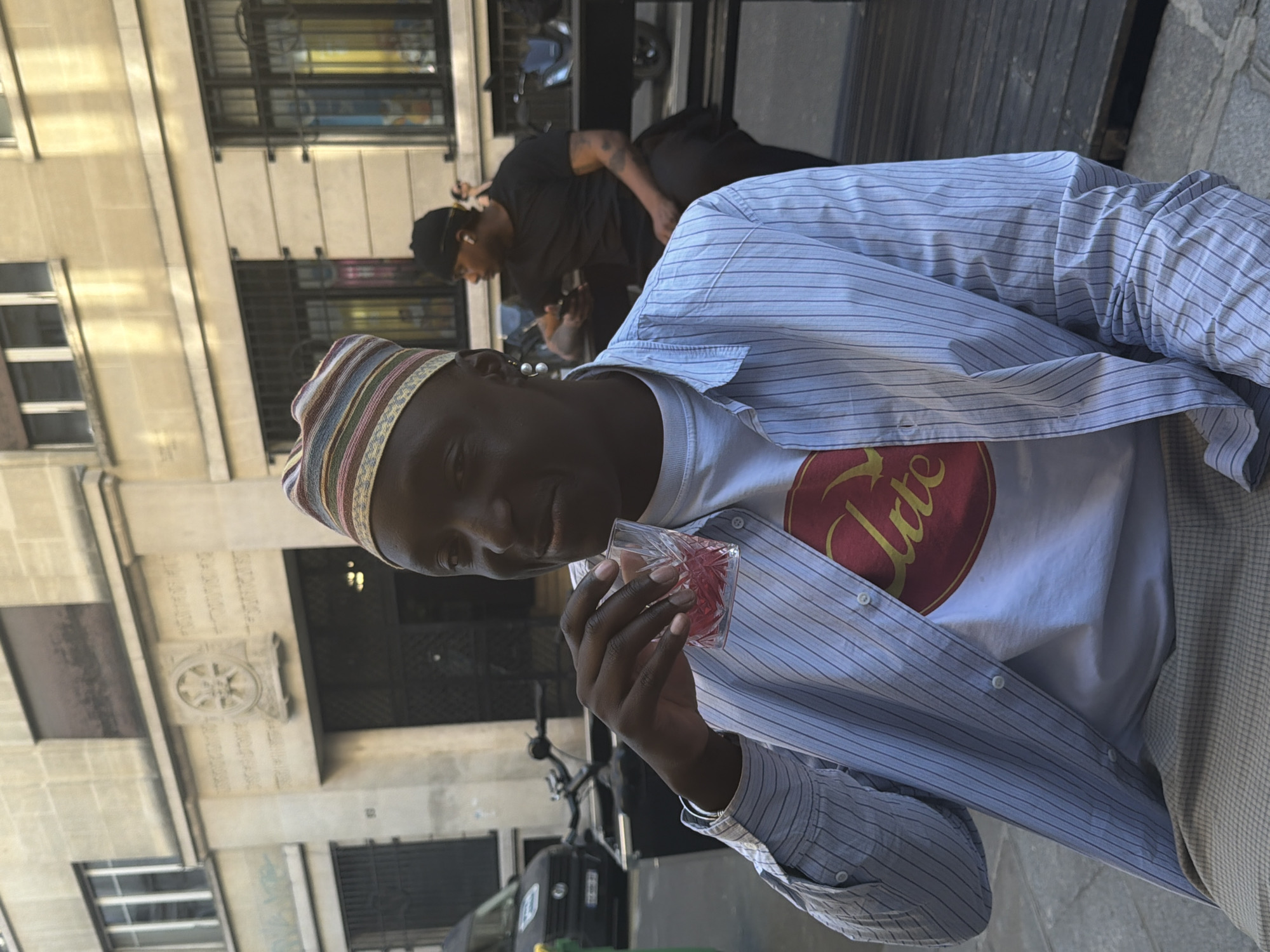 The image captures two African men in Paris, France. The main subject is in the foreground, wearing a multi-colored striped hat, a white T-shirt with a burgundy design, and a light blue striped dress shirt. He is holding and drinking from a glass of red liquid. In the background, another man is seen wearing a black shirt and hat. The architectural backdrop indicates a Parisian street with tall buildings, including a building with an intricate design above the entrance. Parked motorbikes and a car can be seen in the vicinity. The lighting suggests it's daytime, with the scene bathed in sunlight.