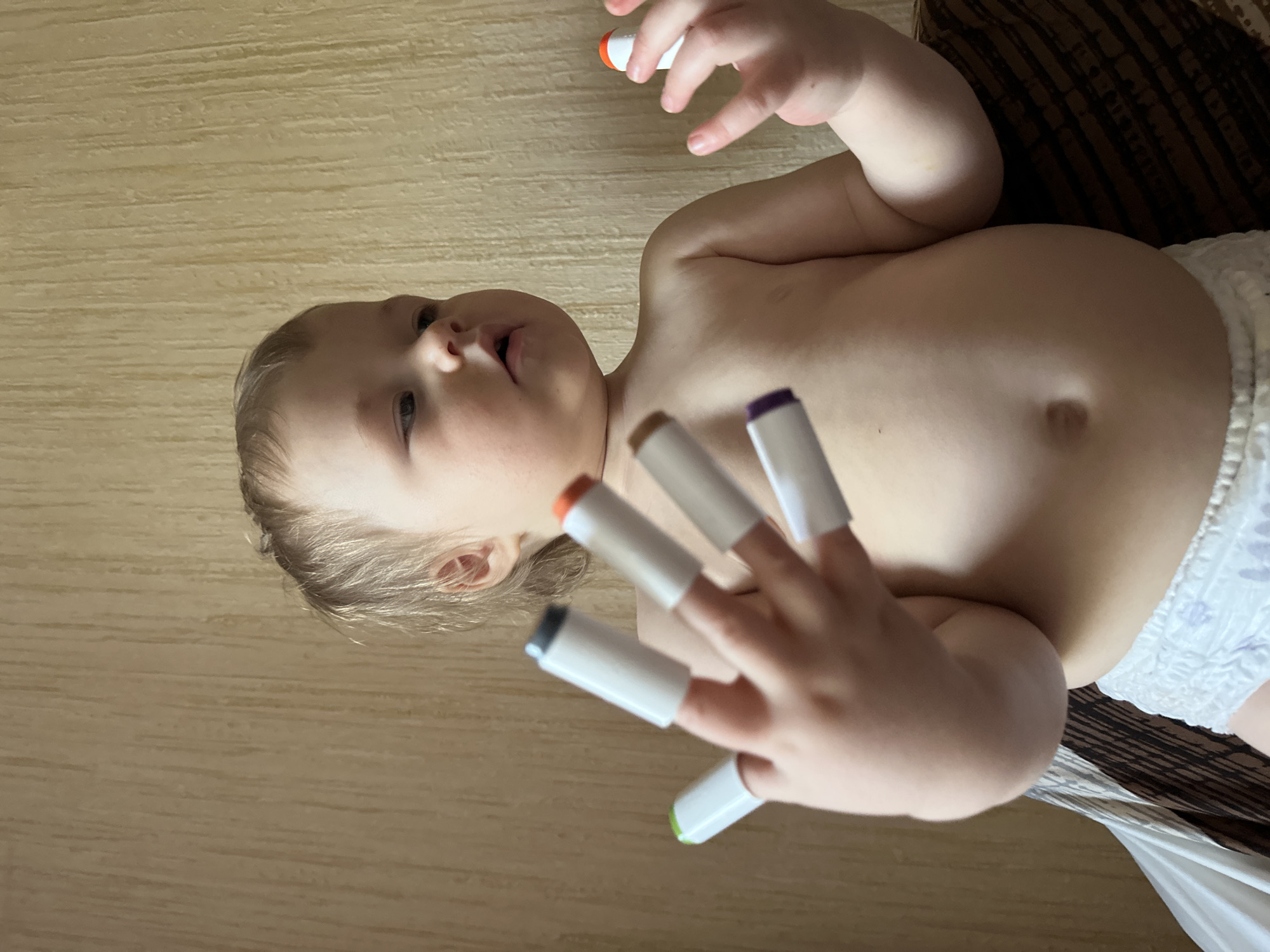A baby lies on its back on a wooden surface, wearing a diaper. On the fingers of its right hand are placed five markers with different colored caps: gray, orange, brown, purple, and green. The baby's left hand holds another orange-capped marker. The background also features part of a dark brown patterned fabric. No indication is given that this image was taken in Nizhny Novgorod, Russia.