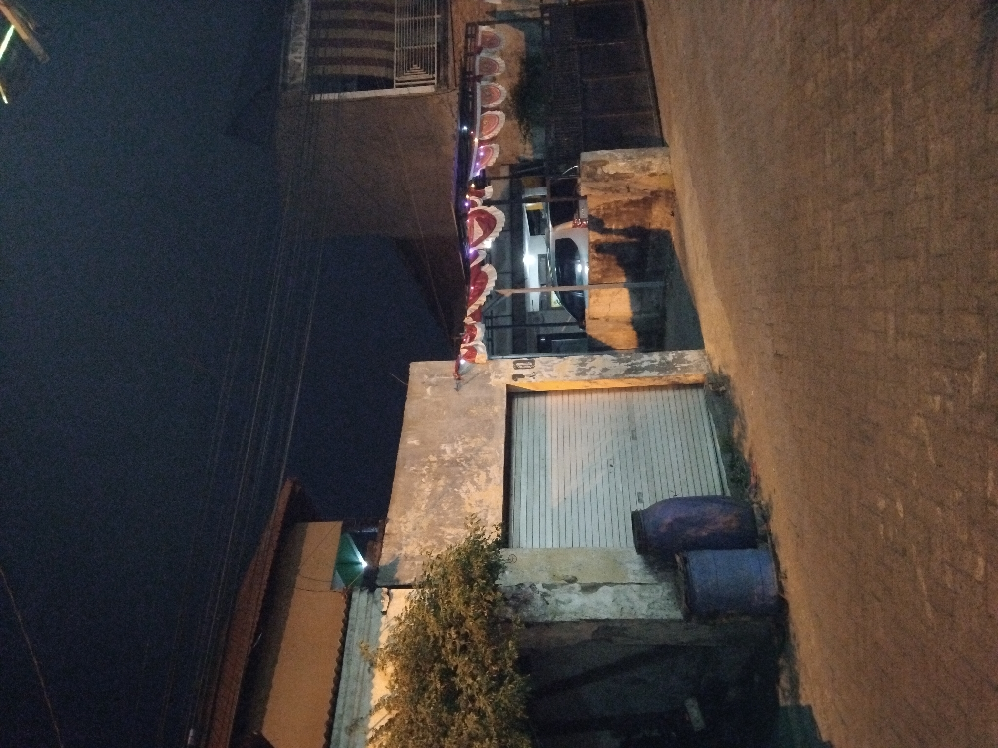 An outdoor, nighttime, low-angle shot captures a street scene in Kepuh Kiriman, Indonesia. A paved road with a brick pattern leads towards a building with a corrugated metal garage door on the left. Two large blue plastic barrels are situated in front of the garage. To the right, a white car is visible parked under a canopy adorned with red and white draped fabric and string lights. The canopy extends over an area with a dark metal gate. The buildings in the background are dimly lit. Overhead power lines are visible against the dark night sky.