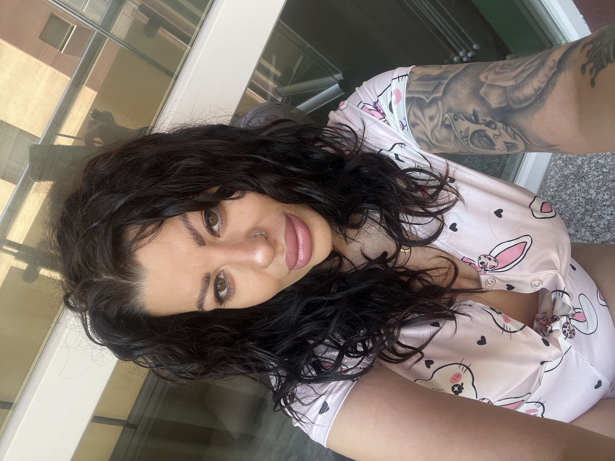 A woman with dark, curly hair is taking a selfie. She has light brown eyes, a nose piercing, and is wearing light pink pajamas with a cartoon bunny print. She is indoors, near a window or balcony overlooking an urban setting with buildings visible in the background. The location is unknown and cannot be determined from the image.