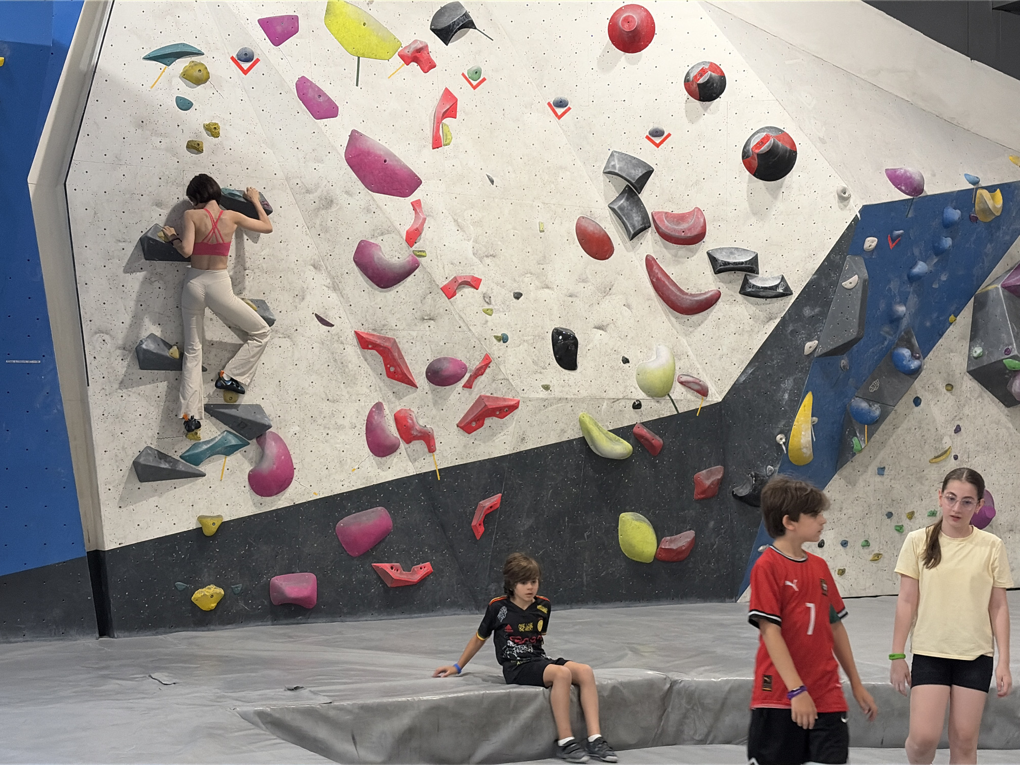 The image shows an indoor climbing gym with a large bouldering wall. A woman in a pink sports bra and beige pants is climbing the wall, gripping a grey handhold. She is positioned in the upper left quadrant of the image. In the lower section of the image, two children are sitting on a mat. A boy in a black soccer jersey and shorts is on the left, and a girl in a yellow t-shirt and black shorts is on the right. The climbing wall has various colored holds, including pink, yellow, red, and blue, with different shapes and sizes. The environment appears to be a recreational facility, likely a climbing gym. The image does not provide any information that suggests the location is Moscow, Russia.