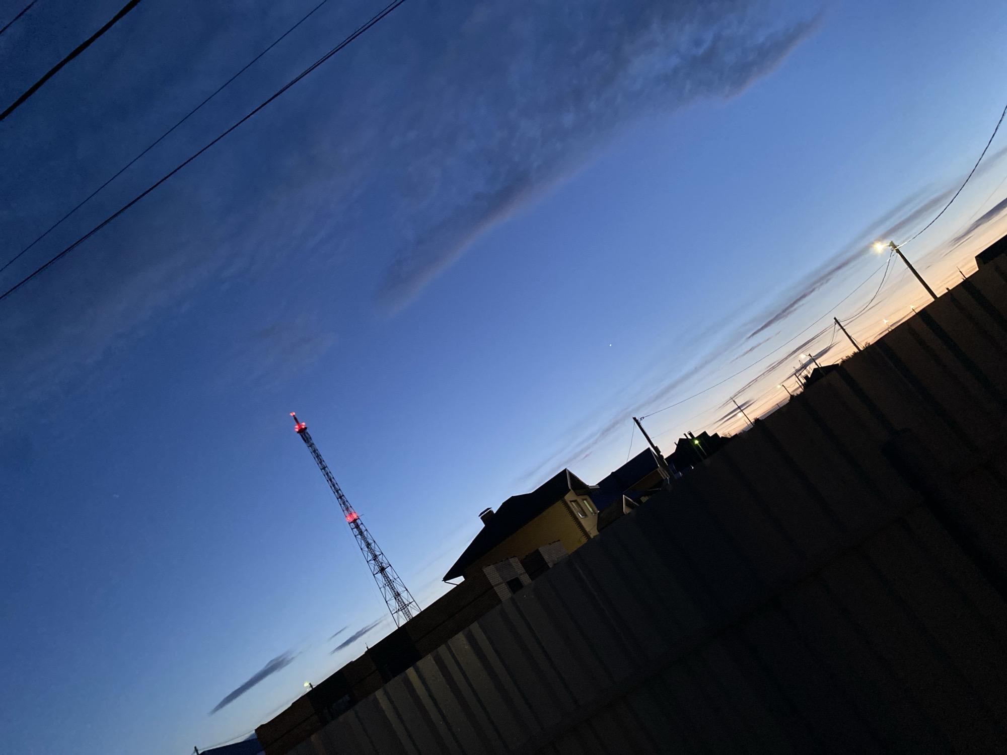 This low-light photograph captures an outdoor scene in Chaikovsky, Russia, during dusk or early night. The sky, a dominant feature, displays a gradient from deep blue in the upper left to a soft, warm orange-yellow closer to the horizon on the right, indicative of twilight. Streaks of thin, elongated clouds are visible across the sky, with utility wires cutting diagonally through the upper left.
A tall, slender metal lattice tower, likely a radio or telecommunications tower, stands prominently on the left side, illuminated by bright red blinking lights at its top. On the right, partially visible behind a dark, corrugated metal fence that diagonally spans the lower portion of the frame, is a house or building featuring a dark roof and lighter-colored walls. Further to the right, a street light on a pole emits a distinct glow against the dimming sky. A small, bright point of light, possibly a star or planet, is subtly present in the sky towards the center-right. The dark fence occupies a significant part of the foreground, creating a strong diagonal compositional line. There is no visible human activity or text in the image, conveying a tranquil and still atmosphere.