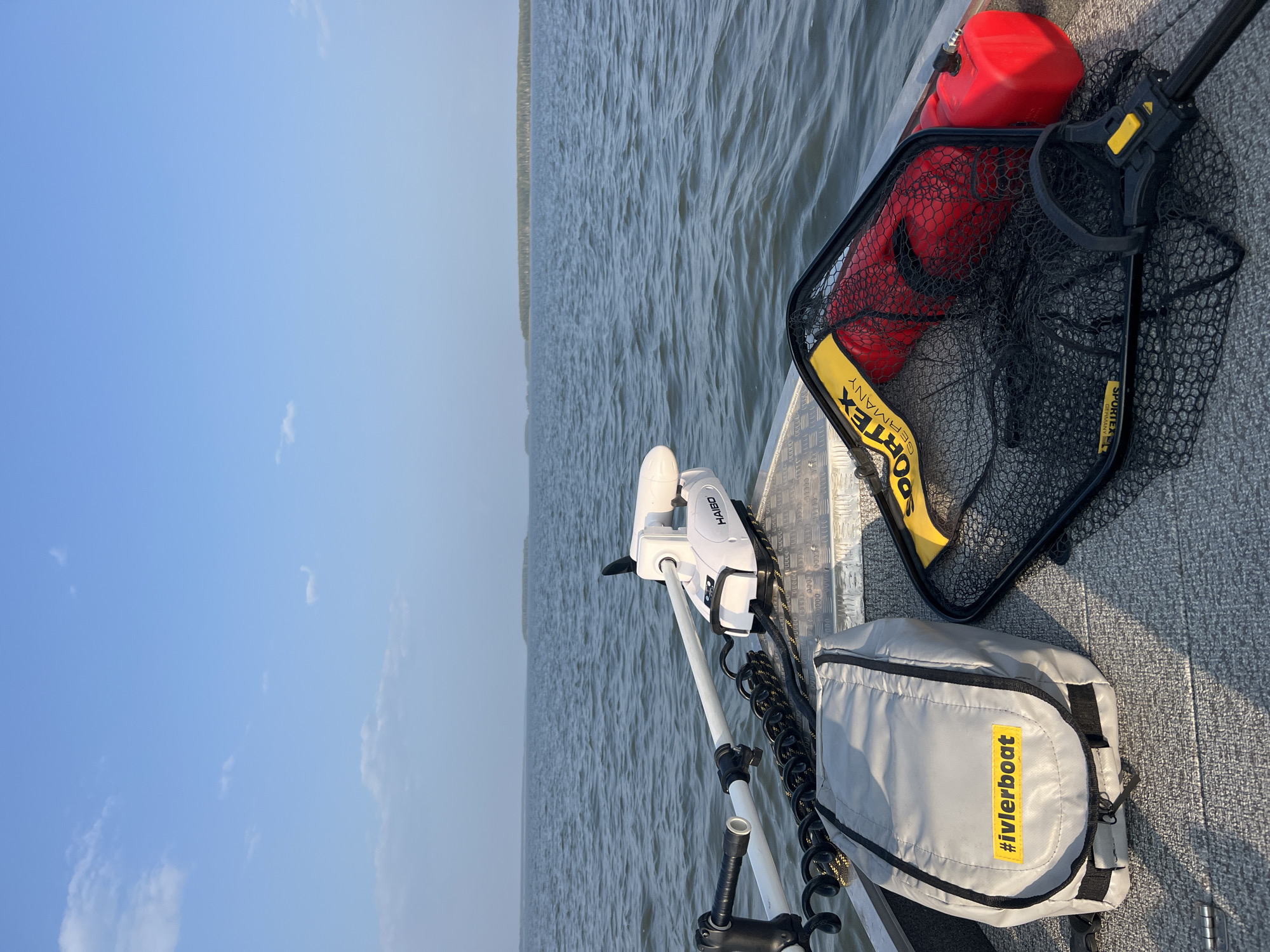 This is a forward-facing, eye-level, medium shot of a boat on the water, possibly in Novopichugovo, Russia, given the text on the bag. The boat is a gray aluminum fishing vessel. In the foreground, on the deck of the boat, are several items: a light gray bag with a yellow rectangular sticker that reads "#ivlerboat", a black fishing net with a yellow and black handle and yellow trim, and a red gas can. The fishing net is partially covering a red float. Behind these items, a white electric trolling motor, branded "HAIBO", is mounted on the front of the boat. The boat is moving across a body of calm water, with a distant shoreline visible on the horizon. The sky is a clear light blue with a few wispy white clouds. The overall scene depicts a peaceful day on the water, likely for fishing.