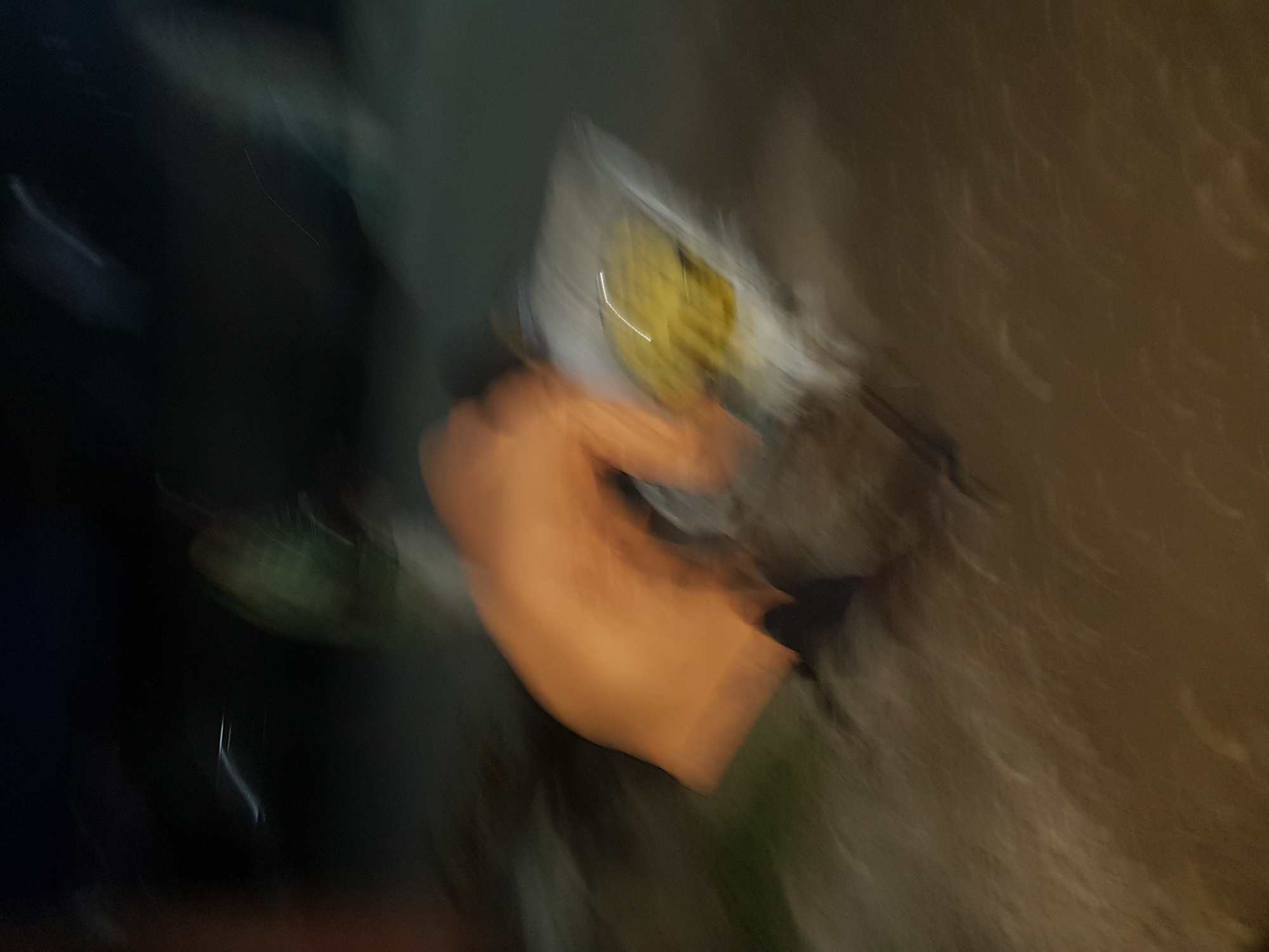 The image is a blurry photograph, likely taken in motion, with unclear details of the scene. There appears to be a person wearing an orange top in the center of the image. To the left is a darker area, possibly representing shadows or objects. The upper-middle area seems to contain a yellow object or pattern on a white surface, which could be a building or a sign. To the right, there are blurry light brown tones, potentially indicating a wall or another structure. The scene is presumed to be in Shangisha, Nigeria, but there are no distinctive visual elements to confirm the location.