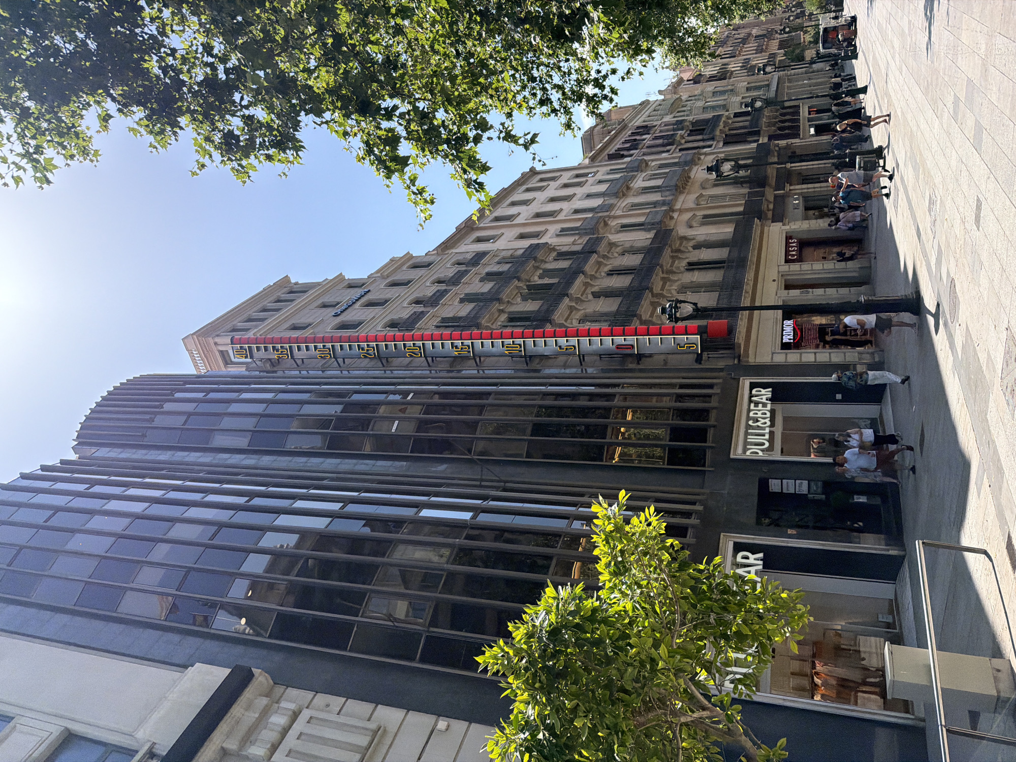 A bright, sunny daytime street view in Barcelona, Spain, features a sidewalk lined with shops and apartment buildings. Dominating the left side of the image is a modern, dark glass-fronted building with vertical silver trim. Adjacent to it, a taller, older building displays a large, vertical thermometer with red and yellow markings, indicating a temperature above 30 degrees Celsius, with the mercury line approaching 35. The thermometer is mounted on a light-colored building with ornate architectural details and balconies.
Several people are walking along the pedestrianized sidewalk. In front of the dark glass building, two storefronts are visible, one with "PULL&BEAR" signage. Further down the street, other shops include "PRIMOR" and "CASAS." Traditional street lamps with ornate posts are spaced along the sidewalk. Lush green trees with full canopies overhang parts of the street, casting dappled shadows on the ground. The sky is clear and blue, suggesting warm weather. The overall scene depicts a bustling urban environment on a warm day.