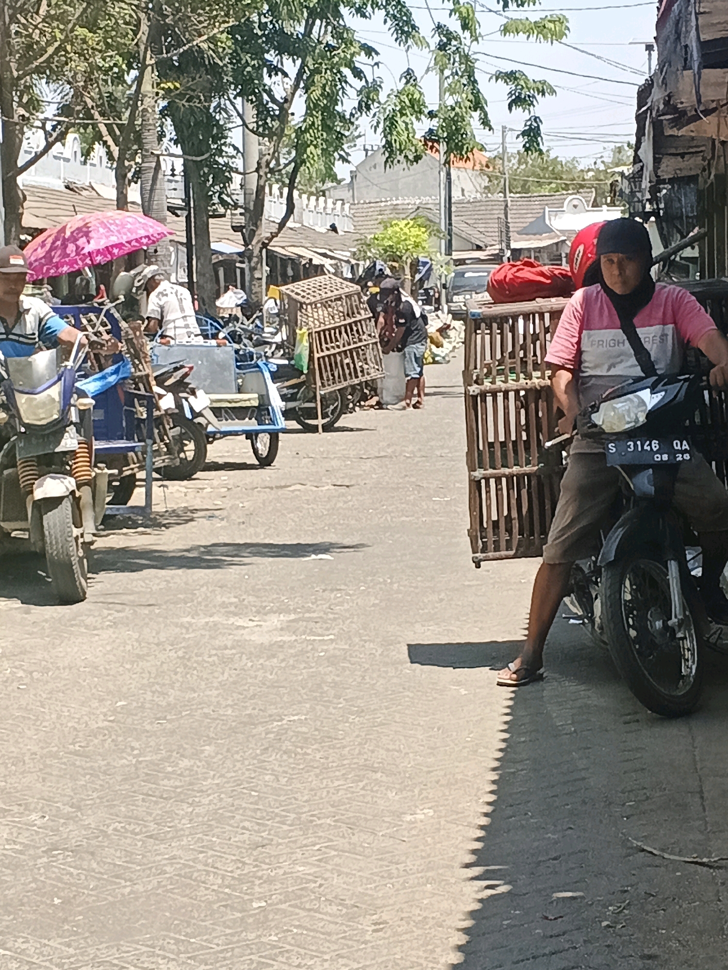 In Krian, Indonesia, a sunny outdoor scene depicts a street lined with buildings and trees. In the foreground, a man wearing a pink shirt and black head covering rides a motorcycle. The motorcycle has a license plate reading "S 3146 QA 08 20". To his left, a wooden cage is visible. Further back, another motorcycle with a blue sidecar is parked, and a person in a blue and white striped shirt is seated on it. A pink umbrella is open nearby. Several other people and various objects, including more wooden cages and red bags, are scattered throughout the background, suggesting a marketplace or bustling street. The ground is paved asphalt.