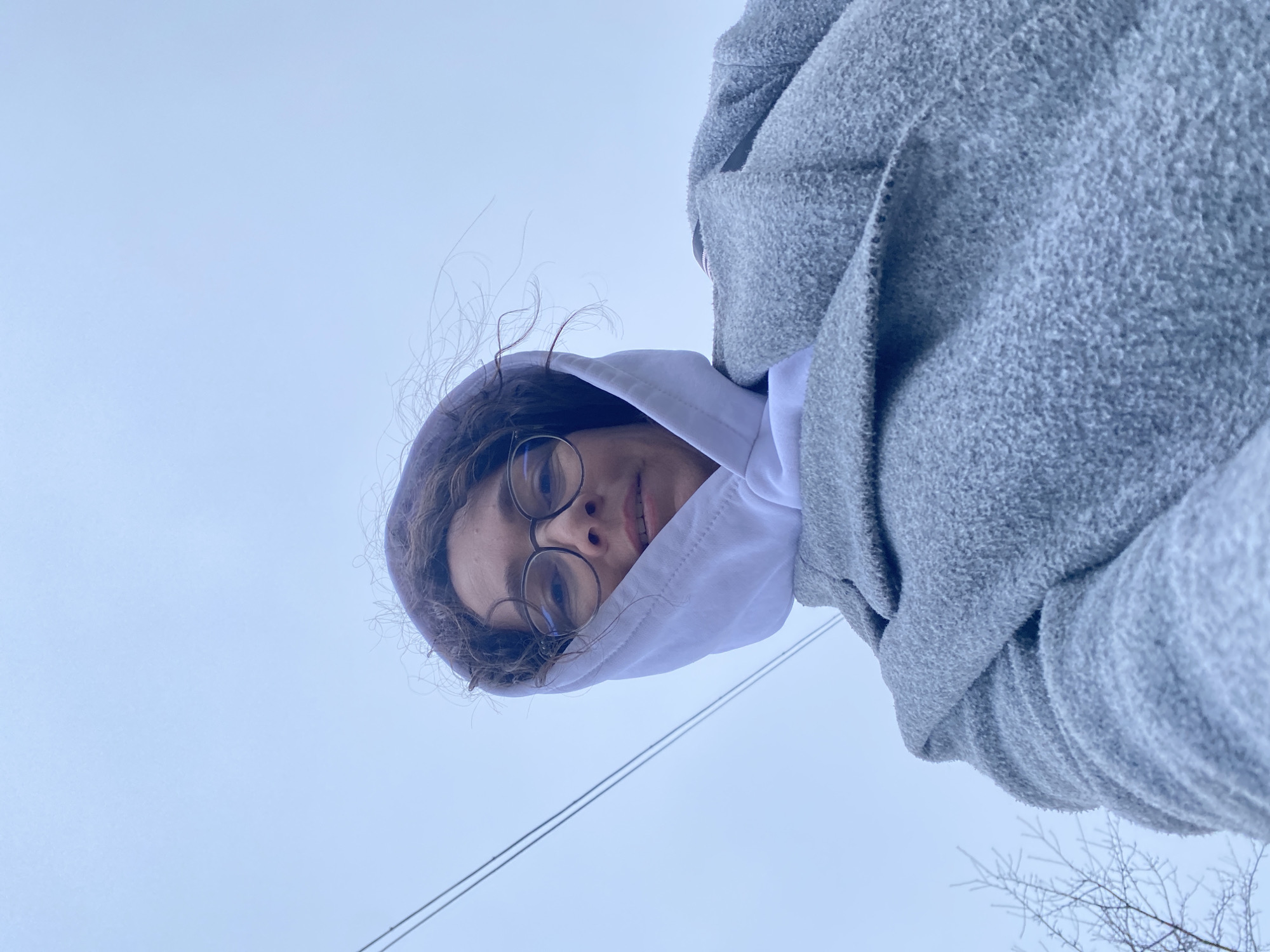A low-angle shot depicts a person wearing glasses, a white hoodie, and a gray fleece jacket. The person is positioned against a pale blue sky, with bare tree branches and overhead power lines visible in the lower right corner of the frame. The location is potentially in or near Прикамский, Russia, based on the user request.