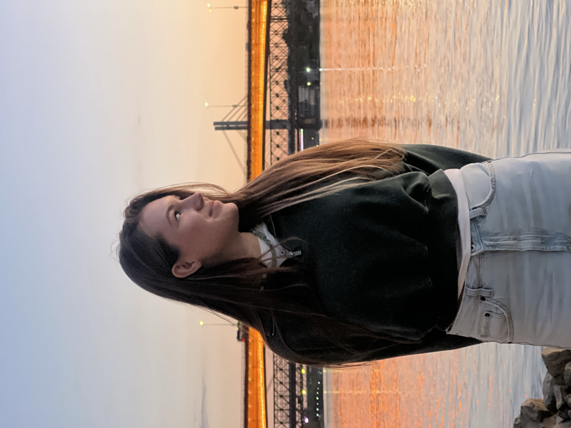 A young woman stands in front of the Sava River in Belgrade, Serbia, with the Branko's Bridge illuminated in the background during sunset. The water reflects the warm light of the setting sun. The woman is wearing a dark green hoodie, a white top, and light blue jeans. She is looking upwards and to the left. She is on a rocky bank.