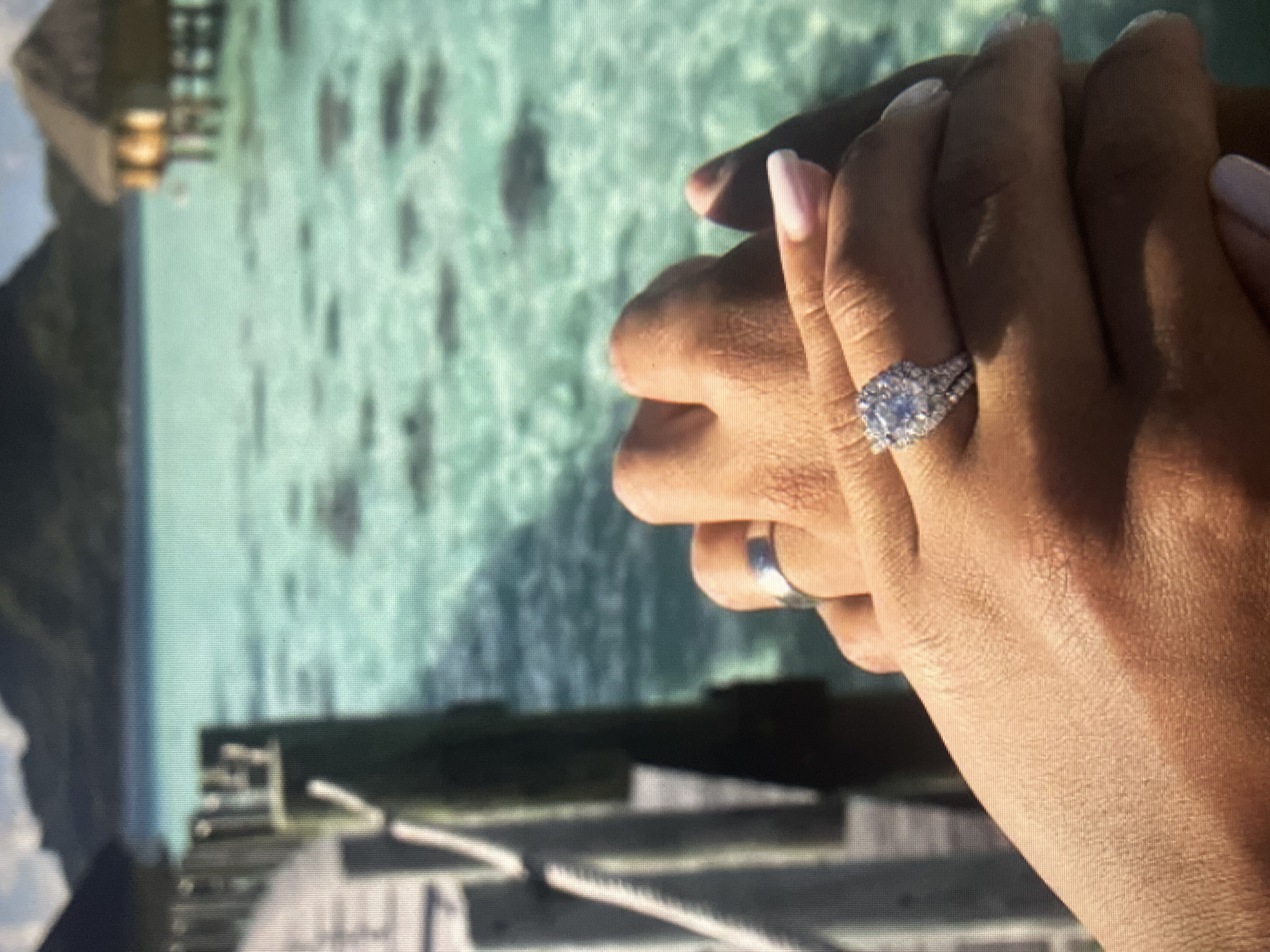 In the foreground, two hands are clasped together, adorned with wedding rings. One hand wears a plain silver band, while the other is embellished with a large, multi-stone engagement ring. The background depicts a serene tropical setting with crystal-clear turquoise water and thatched-roof bungalows on stilts. A wooden pier extends into the water on the left side of the image, and distant mountains form the horizon. There is no indication of the city of Qarağandı, Kazakhstan, in this image.