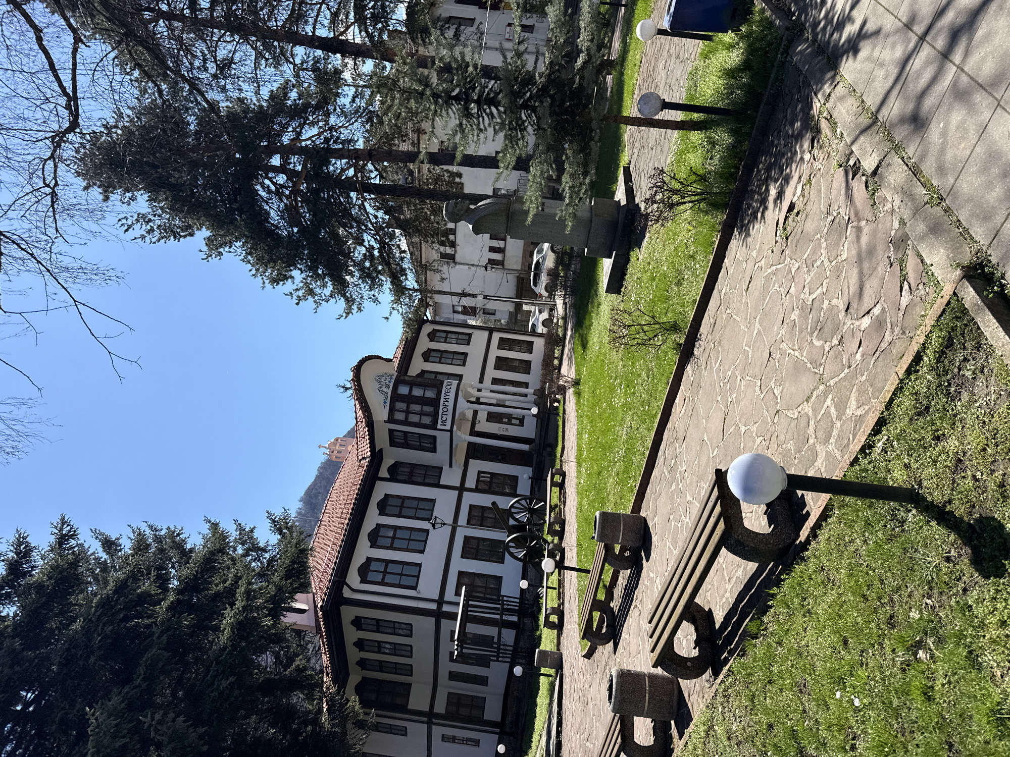 The image depicts the historical museum in Etropole, Bulgaria, on a clear day. The museum is a two-story building with white walls and dark wooden beams, topped with a red-tiled roof. The word "ИСТОРИЧЕСКИ" (historical) is visible above the entrance. In front of the museum, there are several cannons. A paved pathway runs along the side of the museum, with benches and globe-shaped light fixtures. To the left of the museum, lush green trees provide a backdrop. A sculpture can be seen partially obscured by trees in the background.