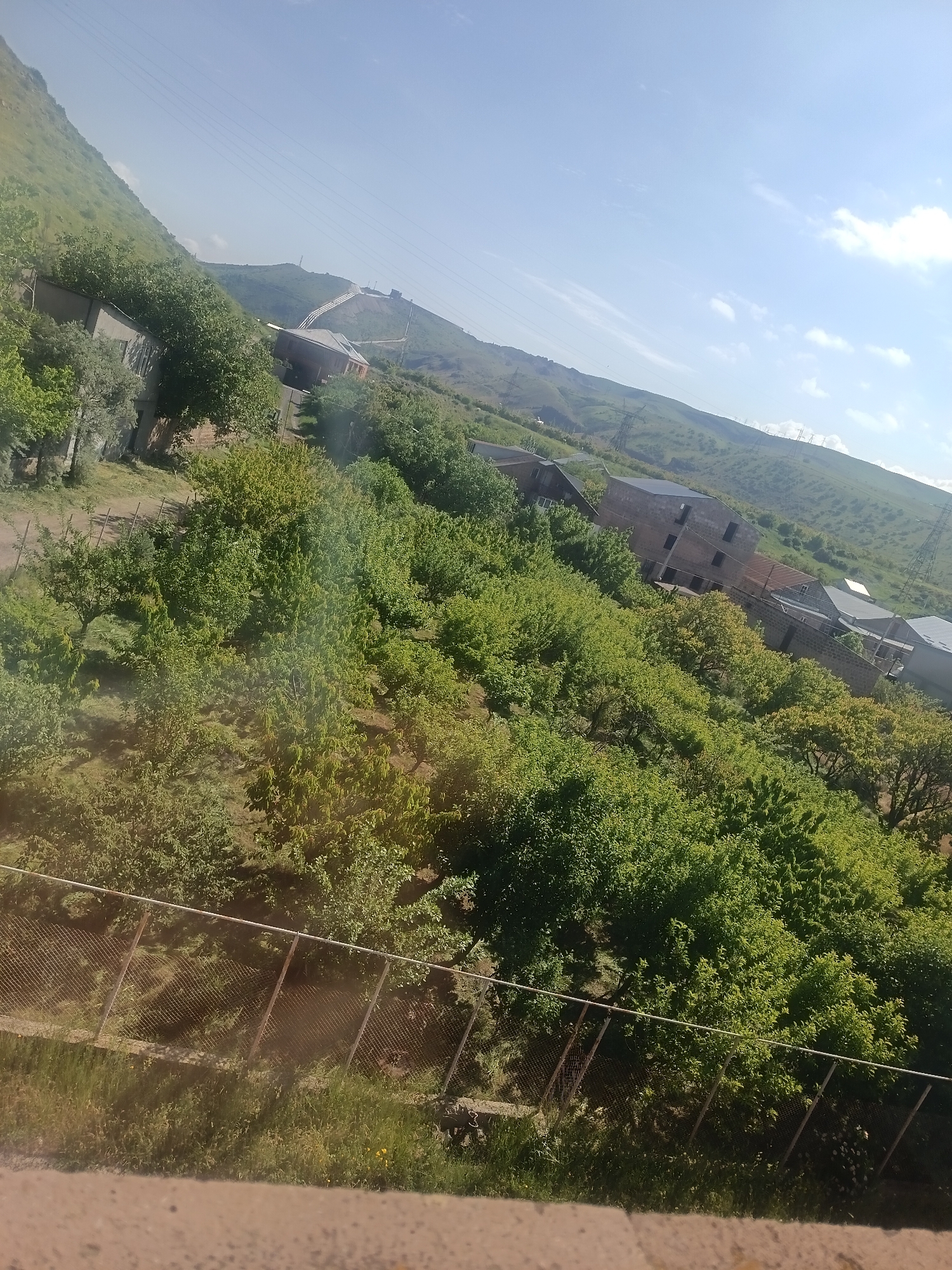 The image depicts a landscape with a prominent hill sloping upwards from the left to the right, covered in greenery and dotted with trees. Several unfinished and completed buildings are scattered across the hillside, some appearing to be multi-story structures. A chain-link fence runs horizontally across the lower part of the frame, separating a grassy area from what appears to be a more overgrown section of land. Power lines and transmission towers are visible on the hill and in the distance, suggesting the presence of infrastructure. The sky is bright blue with scattered clouds. The overall scene suggests a rural or semi-rural setting with ongoing construction and a natural, hilly terrain.
