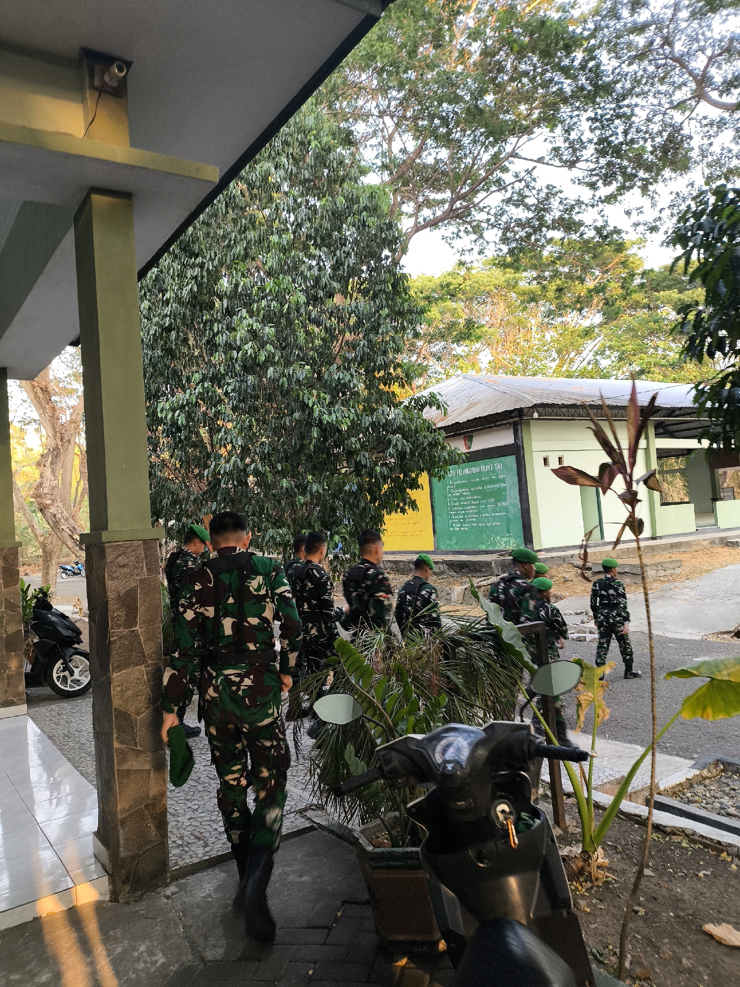 A group of individuals in camouflage military uniforms walks away from the camera, toward a building with a green and white facade. The building has a metal roof and a green wall with writing on it. Trees and foliage are abundant in the background. A motorcycle is parked to the left of the frame, and another motorcycle is partially visible on the right. The ground is paved with interlocking bricks and gravel. The scene appears to be outdoors in a somewhat rural or semi-urban setting. The lighting suggests it is either early morning or late afternoon.