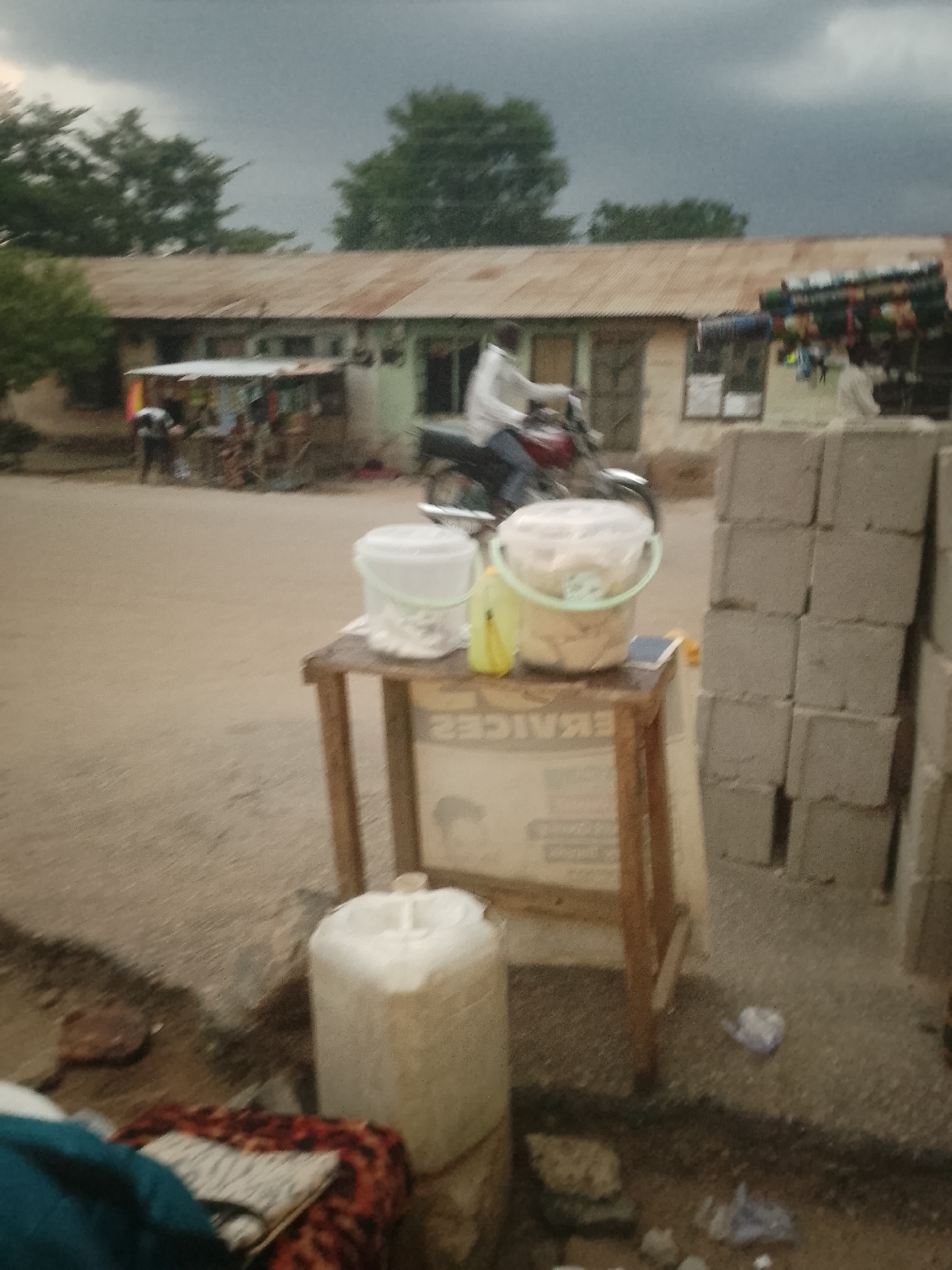 The image depicts a street scene in an unknown location, likely a city. In the foreground, there is a small wooden table supporting two plastic buckets filled with food items, possibly snacks or fried goods. A yellow bottle is also placed on the table. Below the table, a large white container sits on the ground. To the right of the table, a stack of concrete blocks is visible.
In the background, a man is riding a motorcycle on a dirt road in front of a row of single-story buildings with corrugated metal roofs. People are visible near the buildings. The sky is overcast with dark clouds. The overall scene suggests a roadside market or vendor setup in a semi-urban or rural area.