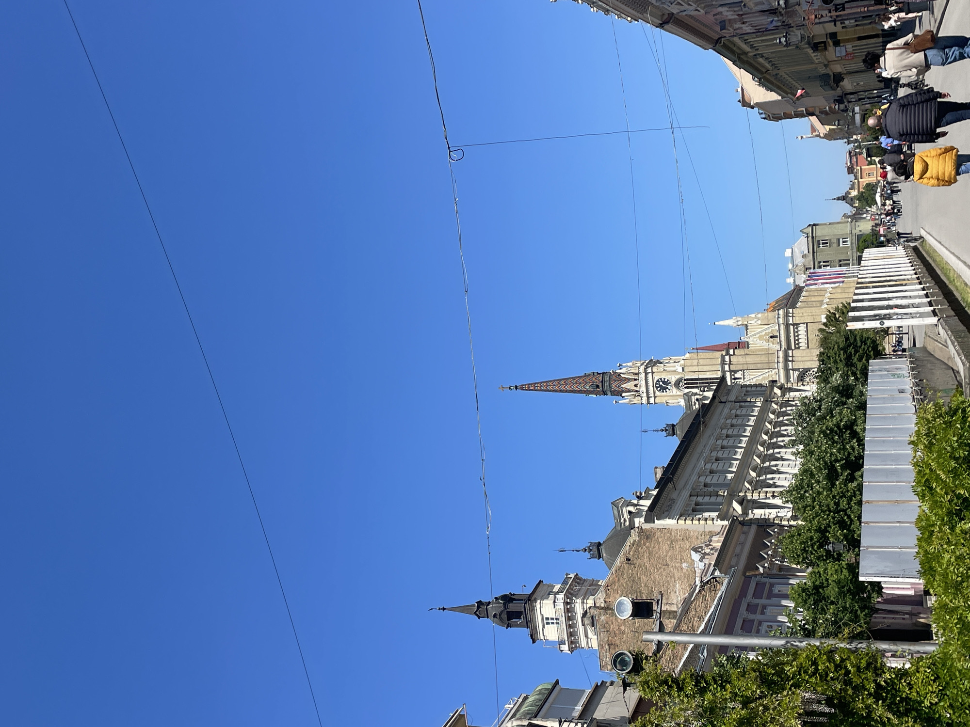 Captured from a street-level perspective, the image presents a sunlit cityscape in Novi Sad, Serbia. The azure sky is crossed by overhead power lines. Dominating the view are the towers of two prominent structures: the Catholic Church of the Name of Mary, identifiable by its tall, colorful spire and clock, and the building of the Serbian National Theatre, marked by its distinct architectural features. Pedestrians can be seen walking along the street, with details of their clothing and actions visible. Various buildings are present, forming the backdrop of the street, and some are partially obscured by trees. A metal street light pole is also visible, adorned with two circular traffic lights. Metal construction barriers line a portion of the street.