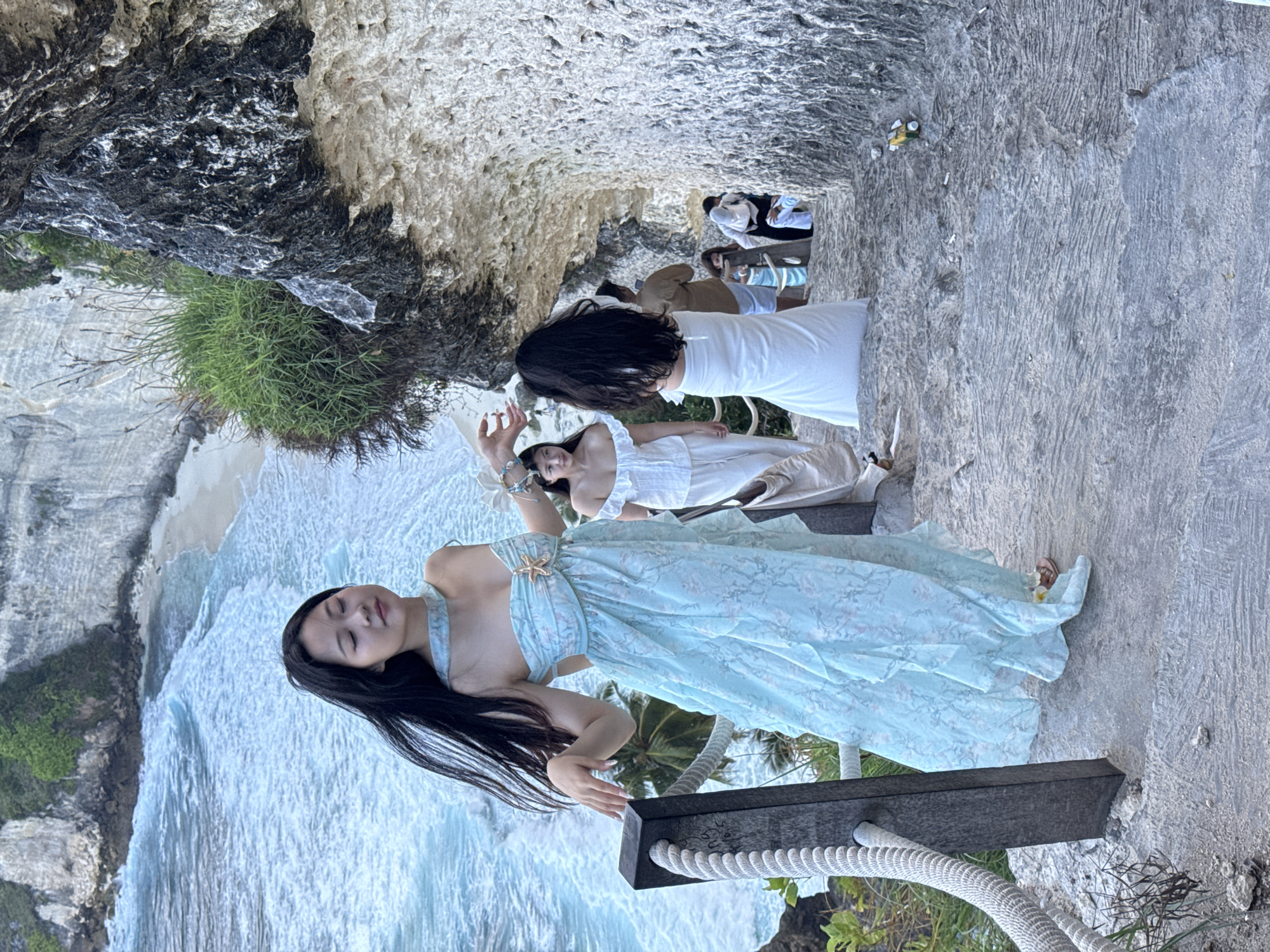 The image depicts a group of people on a stone staircase descending along a cliffside in Nusa Penida, Indonesia. In the foreground, a woman in a light blue, floral-patterned dress with her eyes closed stands on the stairs, her hand resting on a wooden post with a rope railing. Behind her, another woman in a white dress is also on the stairs. Further down the staircase, several other individuals are visible, some walking and others paused. The background reveals a dramatic coastal landscape with turquoise ocean waves crashing against a sandy beach and towering cliffs covered in greenery. The environment is characterized by rugged rock formations and lush vegetation, typical of Nusa Penida's dramatic coastline.
