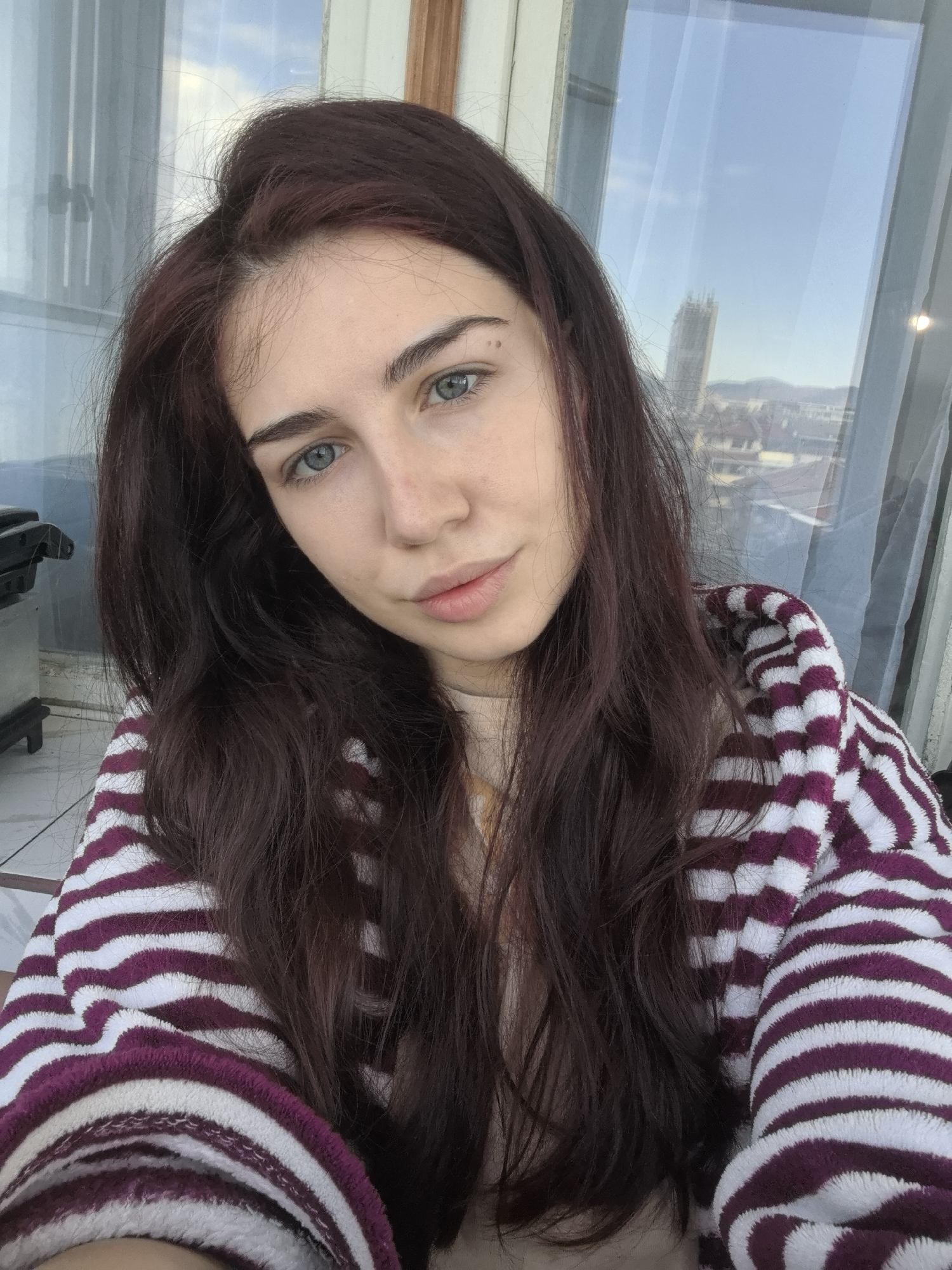 A waist-up selfie shows a person with long, dark auburn hair and blue eyes, wearing a purple and white striped bathrobe. The person faces the camera with a slight, soft expression. Behind them is a large window showing a cityscape under a blue sky with a few clouds. A tall, modern building is visible in the distance. Based on the architecture visible through the window, specifically the tall building in the background, the location appears to be Stara Zagora, Bulgaria. A small, dark object, possibly part of a suitcase or luggage, is visible to the left of the person.