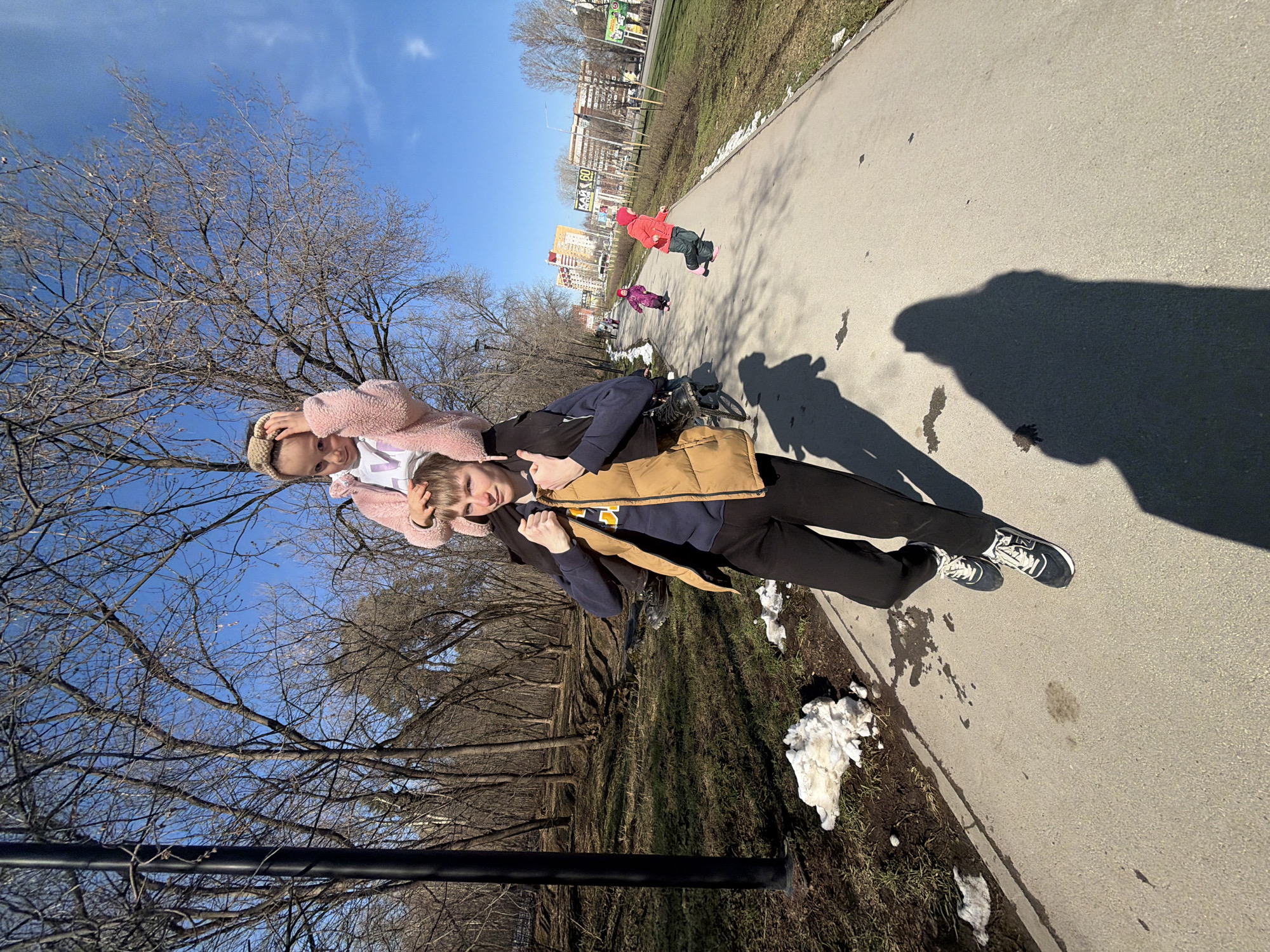 The image shows two people on a sidewalk in what appears to be a park. The park is likely located in Perm, Russia, as buildings characteristic of the city are visible in the background. A child in a red coat, with at least one other child nearby, is visible further down the path. Patches of snow linger on the side of the path. The primary subjects are two people; the person in a tan vest is carrying the other person on their back. The ground is concrete, and the shadow of the figures is visible on the path. Bare trees are present on either side of the path. A metal pole is partially visible in the lower left corner.